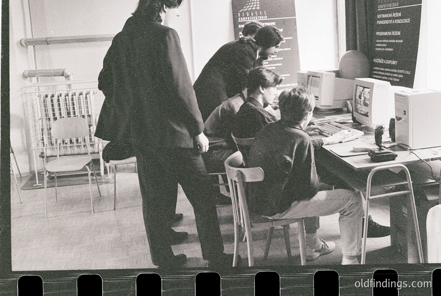 Black-and-white classroom scene featuring early computer education. Three students seated at desks with CRT monitors, likely late 1980s–early 1990s. Teacher in dark coat supervises, while a poster on the wall references "Alcatel" and educational programs. Functional, utilitarian classroom design with wooden chairs and basic desks.
