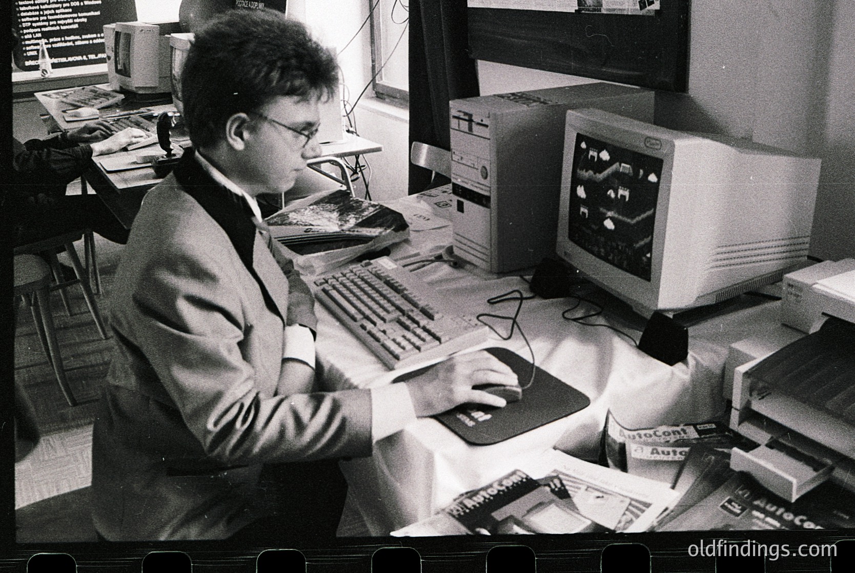 Retro office scene featuring a man in a suit using a vintage **Commodore Amiga** with a monochrome monitor displaying a simple graphic. Surrounding equipment includes a dot-matrix printer, floppy disk stacks, and manuals. Likely late **1980s** workspace.