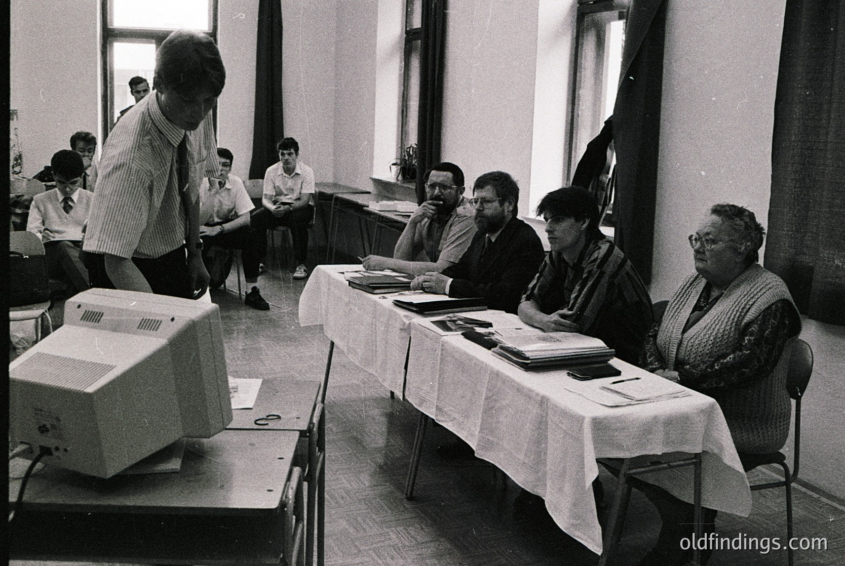 Mid-20th century classroom setting with early computer demo. Man in striped shirt operates a bulky desktop computer (1970s-80s tech) while seated adults observe. Cluttered desks with papers, books, and typewriters reflect institutional learning. Institutional architecture: high ceilings, large windows, and tiered seating.