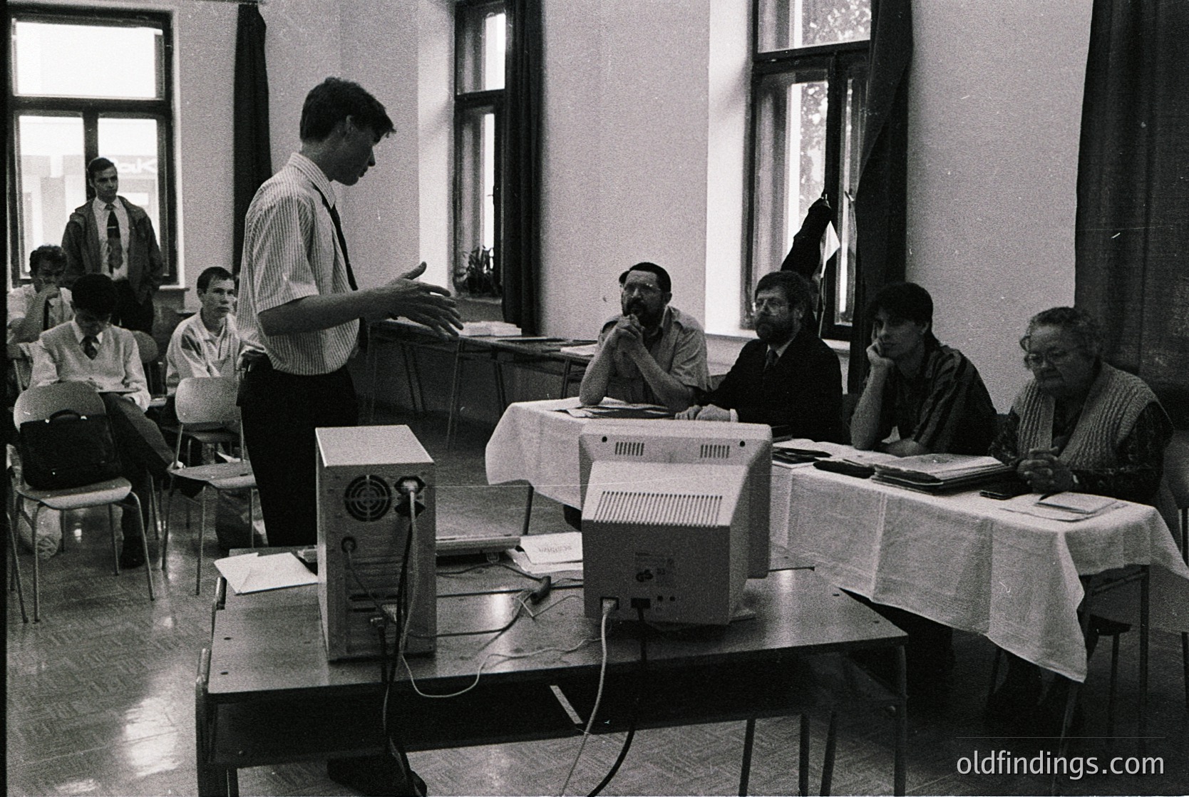 Early 1980s classroom featuring early desktop computing. Instructor demonstrates a bulky CRT-based computer system with external printer, surrounded by seated students engaged in discussion. Wooden desks, white tablecloths, and large windows with blinds. Formal attire suggests institutional setting.