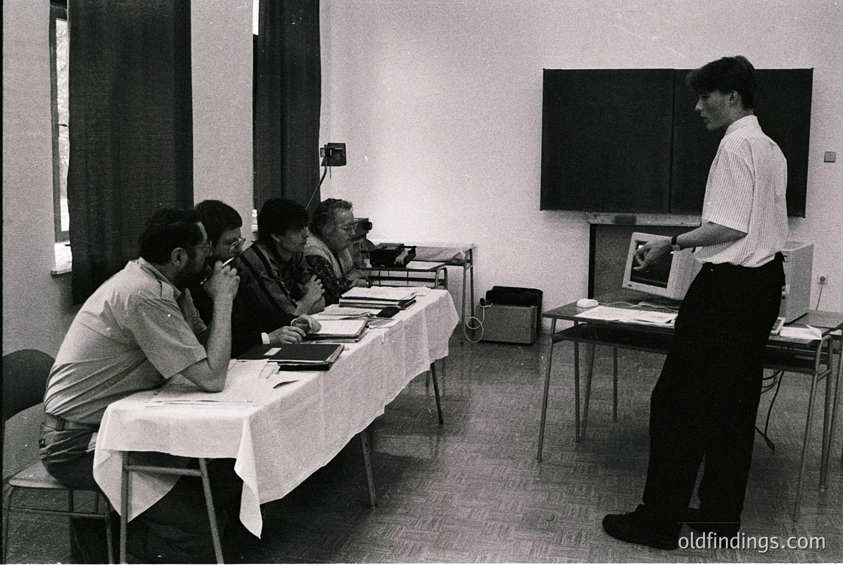 Mid-20th century conference room with four seated attendees reviewing documents at a table, one standing presenter using a projector. Plain white tablecloths, wooden floor, and minimalist decor suggest institutional or academic setting. Likely or .