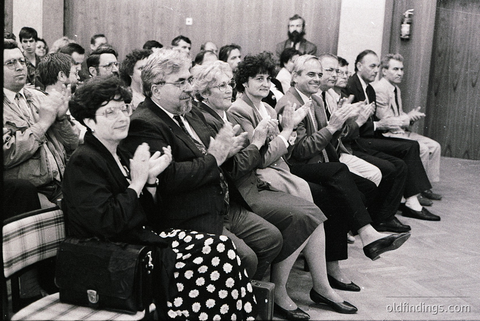 Audience clapping during a formal event in the 1970s, likely a conference or lecture. Indoor setting with wooden chairs and a plain wall. Attendees wear business attire—men in suits, women in dresses or skirts. One woman in a polka-dot skirt stands out. Crowded, attentive atmosphere.