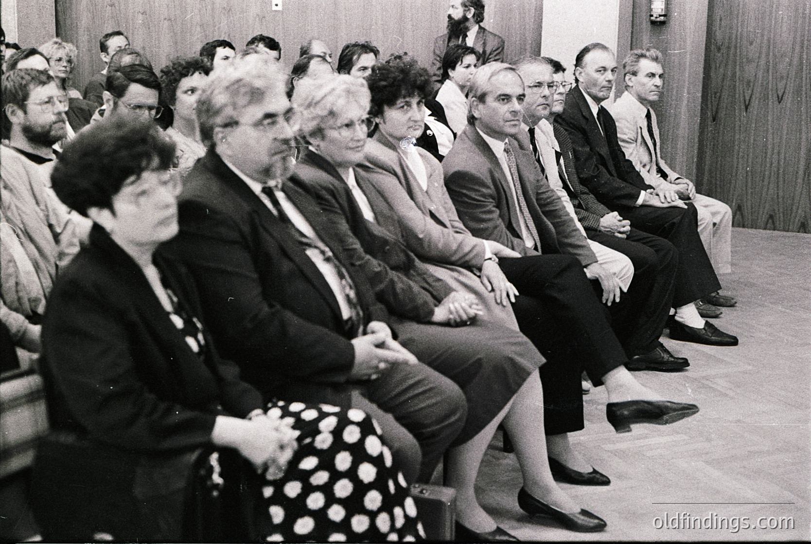 A formal indoor gathering of adults seated in rows, likely a conference or meeting in the 1970s–1980s. Wood-paneled walls and simple wooden chairs suggest institutional or corporate setting. Attendees wear business attire (suits, blazers), indicating professional context. Black-and-white composition highlights mid-century design.
