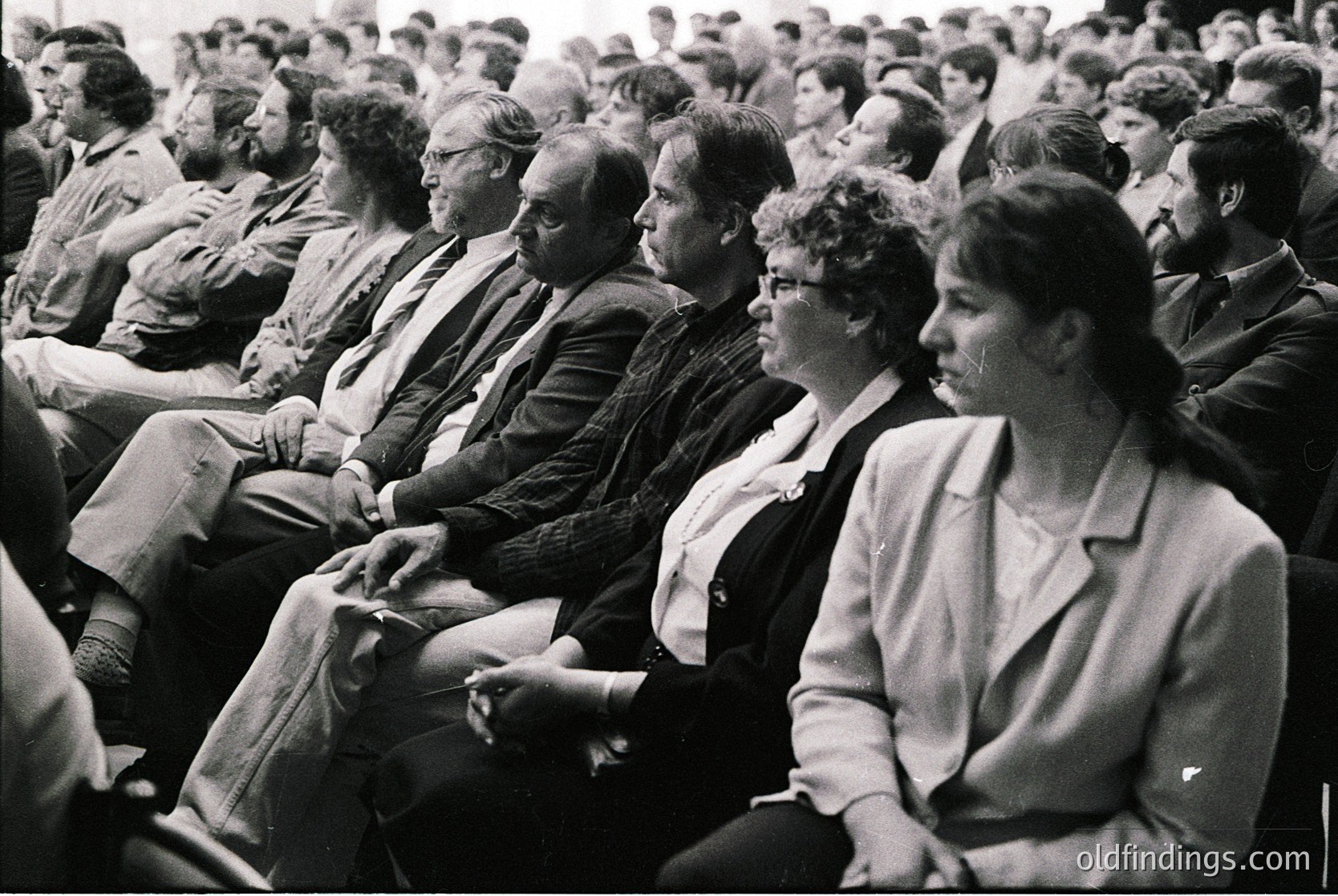 Black-and-white audience of seated adults in formal attire, likely attending a public event or lecture. Men wear suits, ties, and some sport short hair; women in blazers and dresses. Mid-20th century (1950s–1970s) setting, possibly institutional or cultural venue.