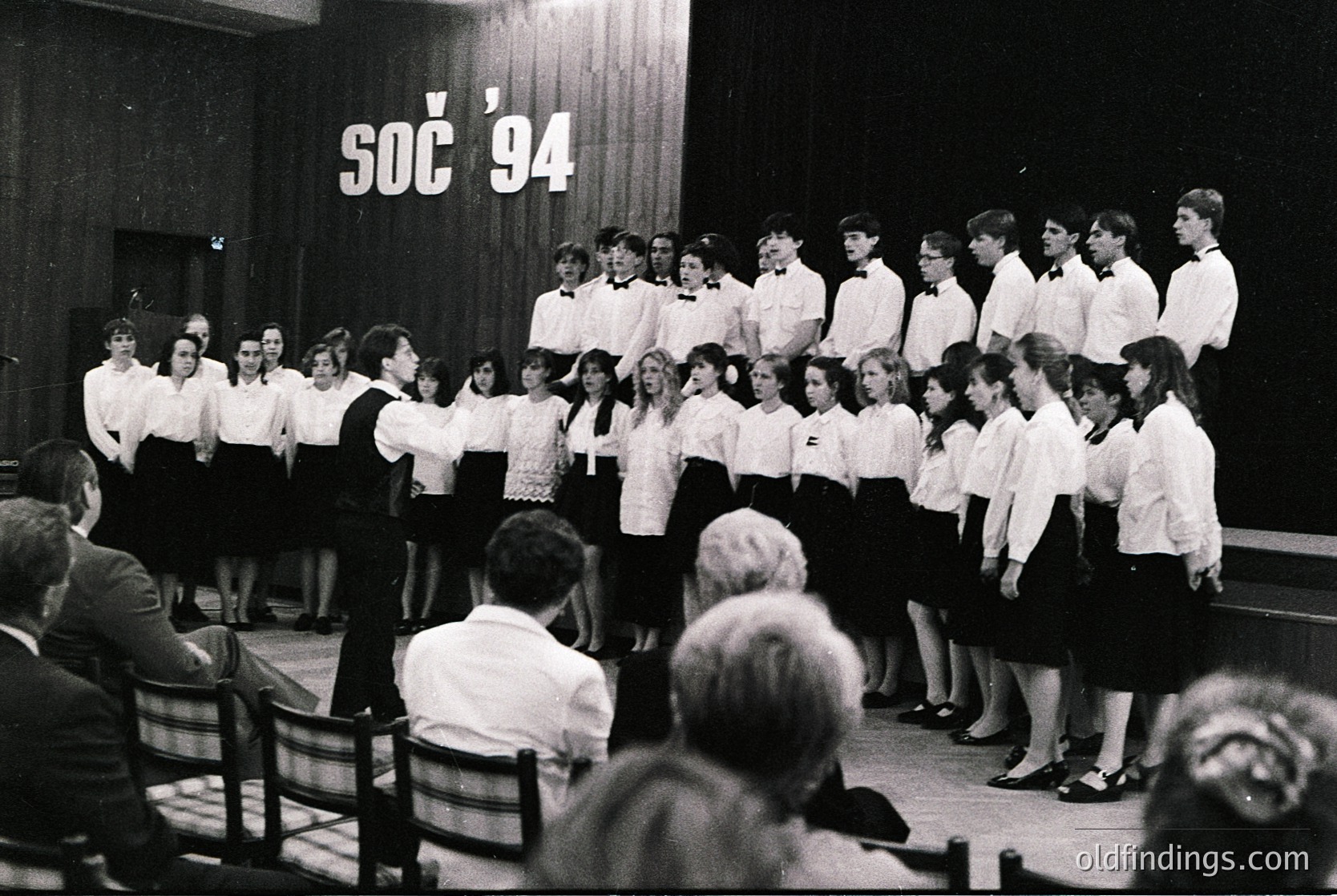 Youth choir performing in a Soviet-era indoor venue, 1970s–1980s. Uniform white shirts with black vests, led by a conductor. Sign "SOČ '94" suggests a Soviet Youth Organization event. Wooden chairs and stage lighting indicate a formal, institutional setting. Č