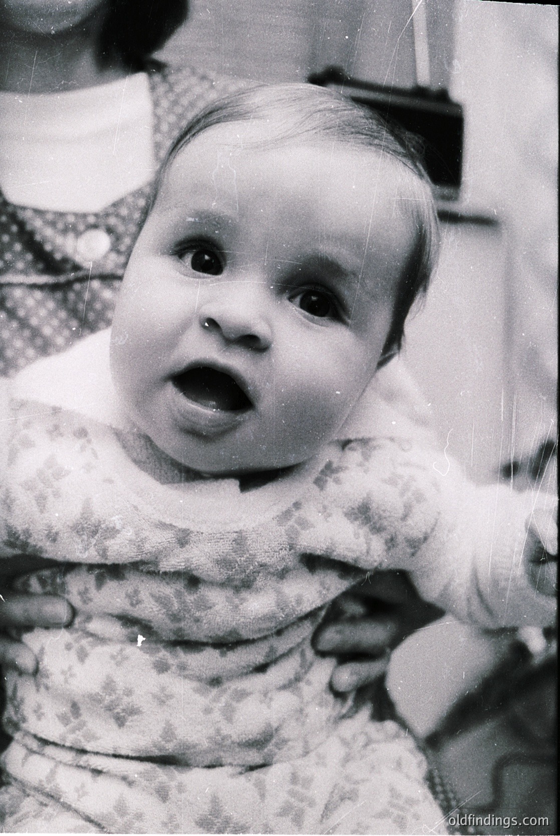 Mid-20th century black-and-white portrait of an infant in a floral-patterned dress, held close by an adult’s hands. The child’s surprised expression and soft focus evoke nostalgia. Likely or family photography.