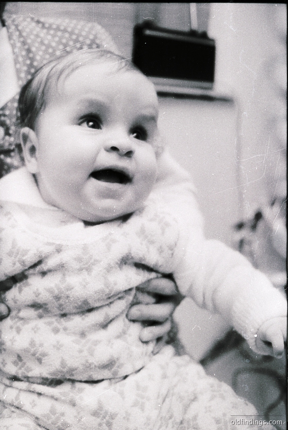 Vintage black-and-white photo of an infant in a knitted sweater, seated indoors with blurred adult hands holding them. Soft focus and grainy texture suggest mid-20th century (1950s–1960s). Warm, candid expression captures innocence.