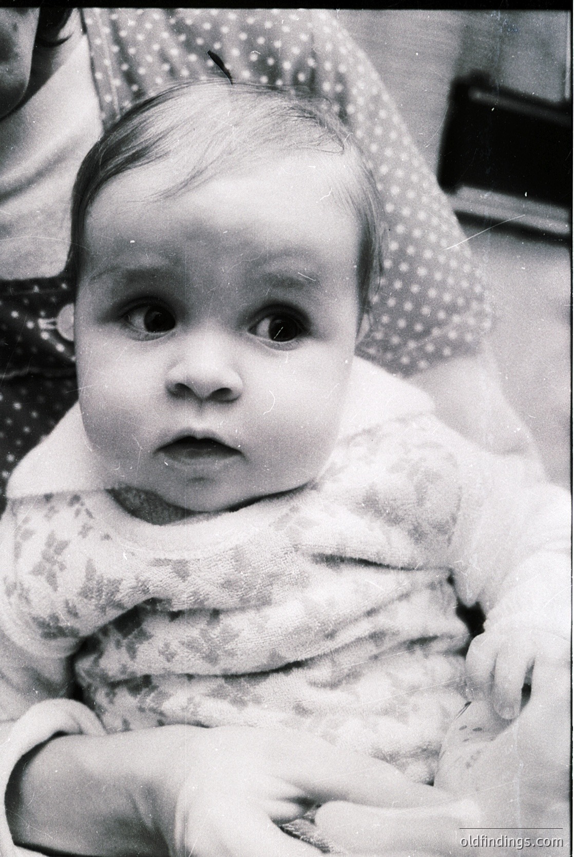 Black-and-white portrait of an infant with direct gaze, dressed in a textured knit garment. Soft focus and vintage framing suggest mid-20th century (1950s–1970s). Warm, candid expression captures innocence and curiosity.