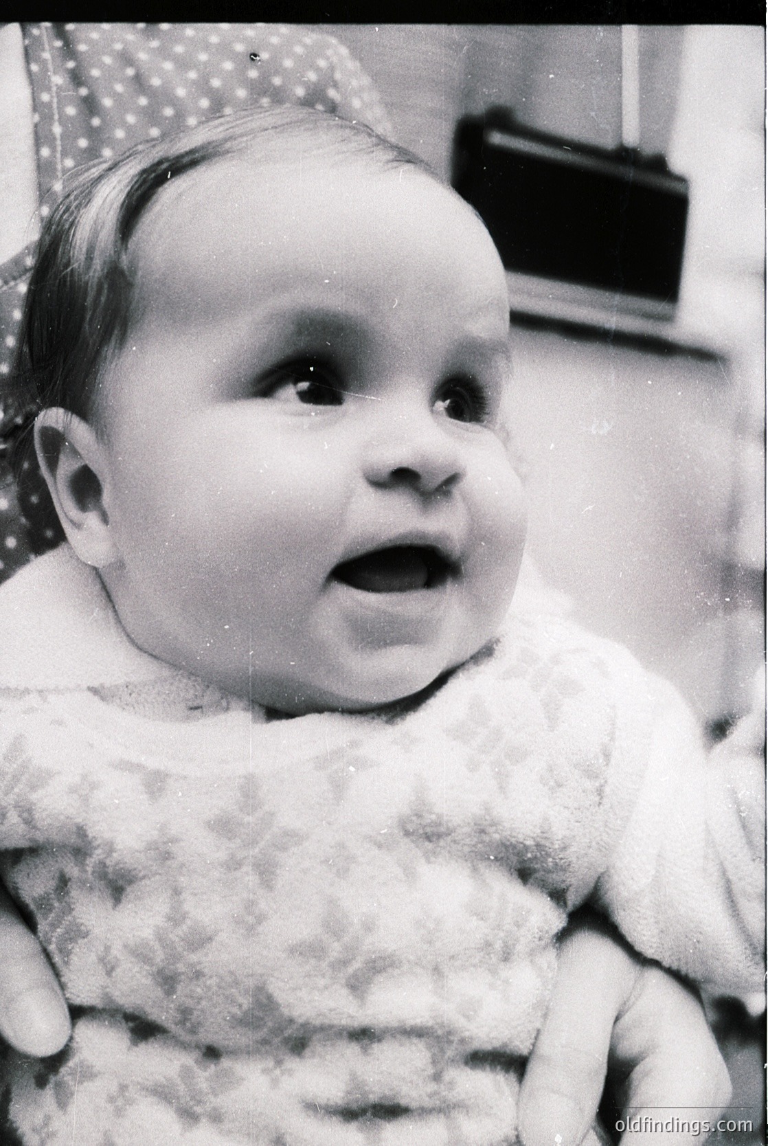 Vintage black-and-white portrait of an infant in a patterned knit garment, seated indoors. The child’s expressive face captures a moment of surprise or curiosity. Background includes a patterned wall and a vintage television set, suggesting mid-20th-century domestic life.