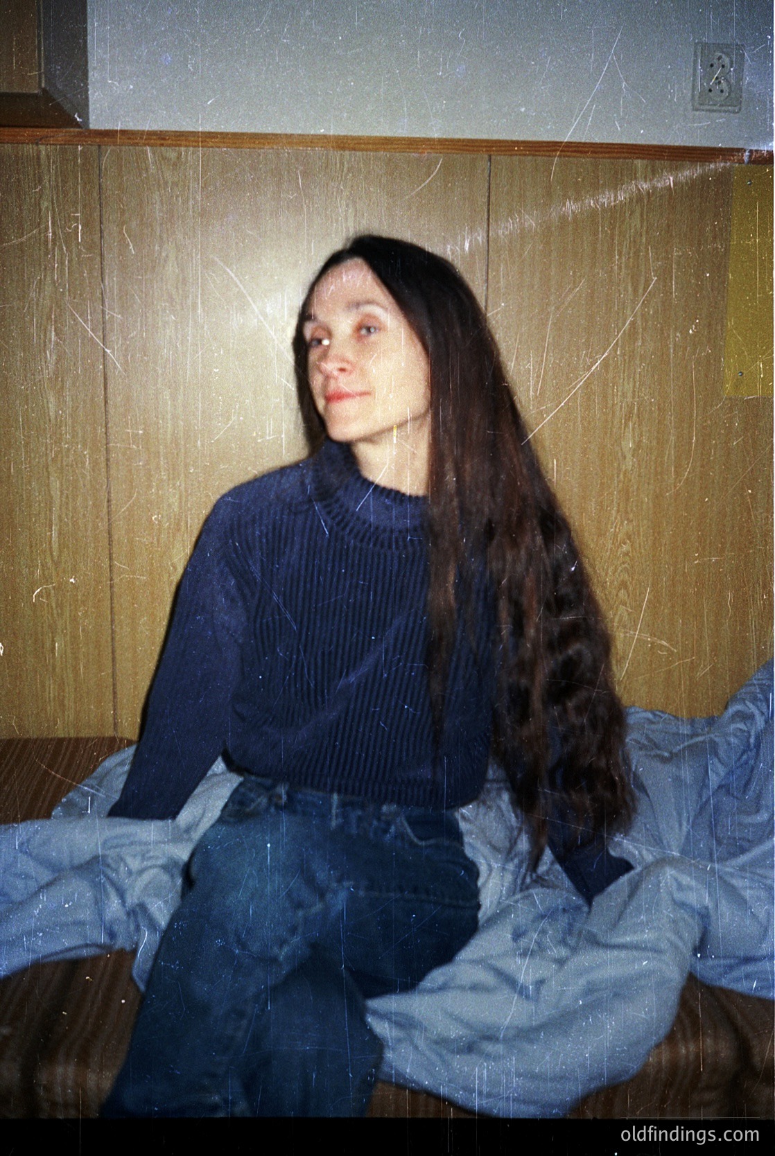 Vintage portrait of a person with long, straight hair in a ribbed sweater and high-waisted jeans, seated on a bed with a striped blanket. Warm, slightly aged lighting suggests indoor domestic setting, likely mid-20th century.