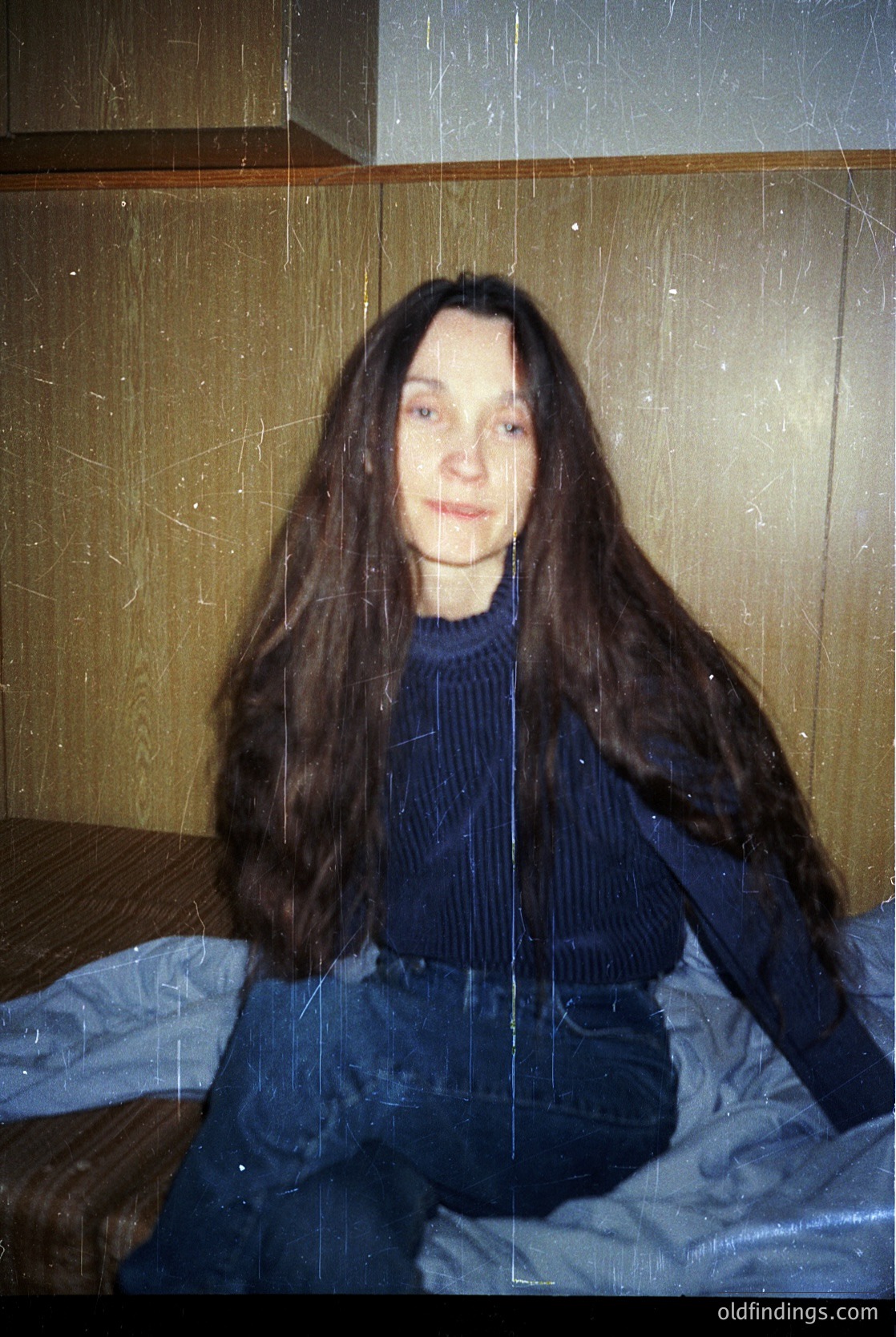 Portrait of a person with long, straight hair in a vintage indoor setting. The individual wears a dark blue ribbed sweater and matching trousers, seated on a bed with a striped blanket. The room’s walls appear textured with peeling wallpaper, suggesting mid-20th-century decor. Soft, diffused lighting enhances the nostalgic atmosphere.