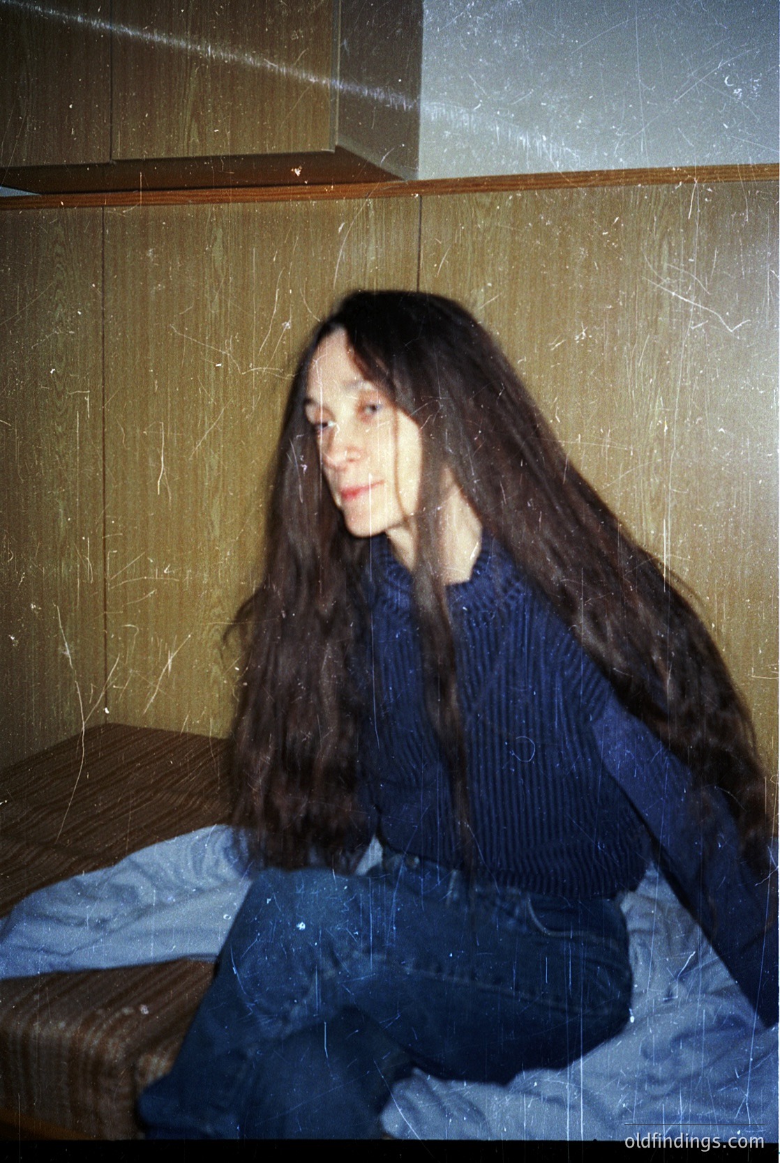 Portrait of a person with long, straight hair in a vintage-style sweater and cropped trousers, seated on a bed with a striped blanket. The room’s peeling wallpaper and wooden headboard suggest mid-20th century domestic interior. Soft, diffused lighting enhances a candid, introspective mood.