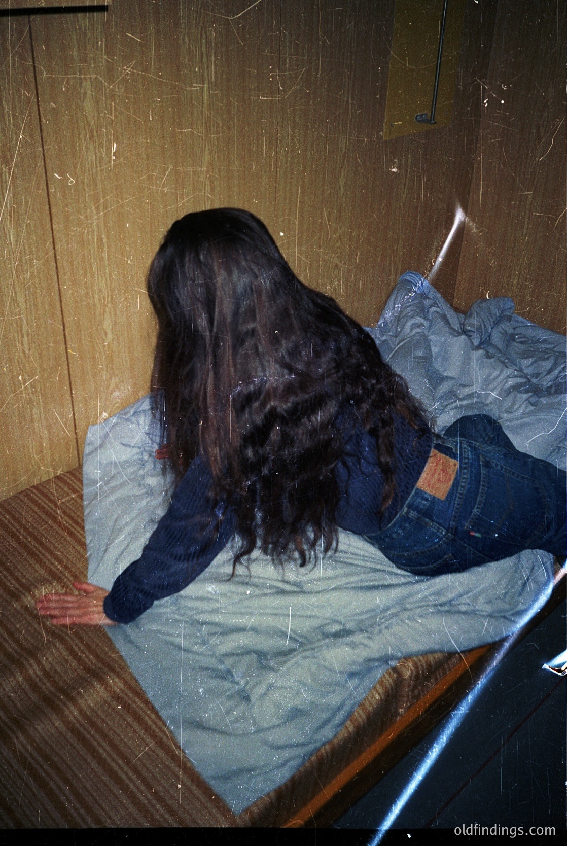 Vintage-style portrait of a person lying on a wooden bed with a faded blue blanket, wearing a dark sweater and distressed jeans with a visible patch. Warm, dim lighting and peeling wallpaper suggest a mid-20th-century interior. #