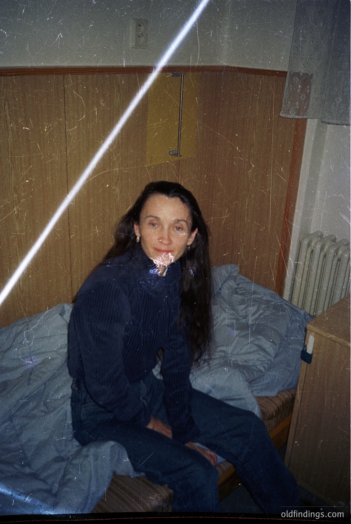 Vintage indoor portrait of a woman seated on a single bed in a utilitarian room, likely from the 1970s–1980s. She wears a dark, textured sweater and jeans, holding a cigarette. The room features wallpaper with a geometric pattern, a wooden-framed mirror, and a radiator. The lighting suggests a single overhead bulb.