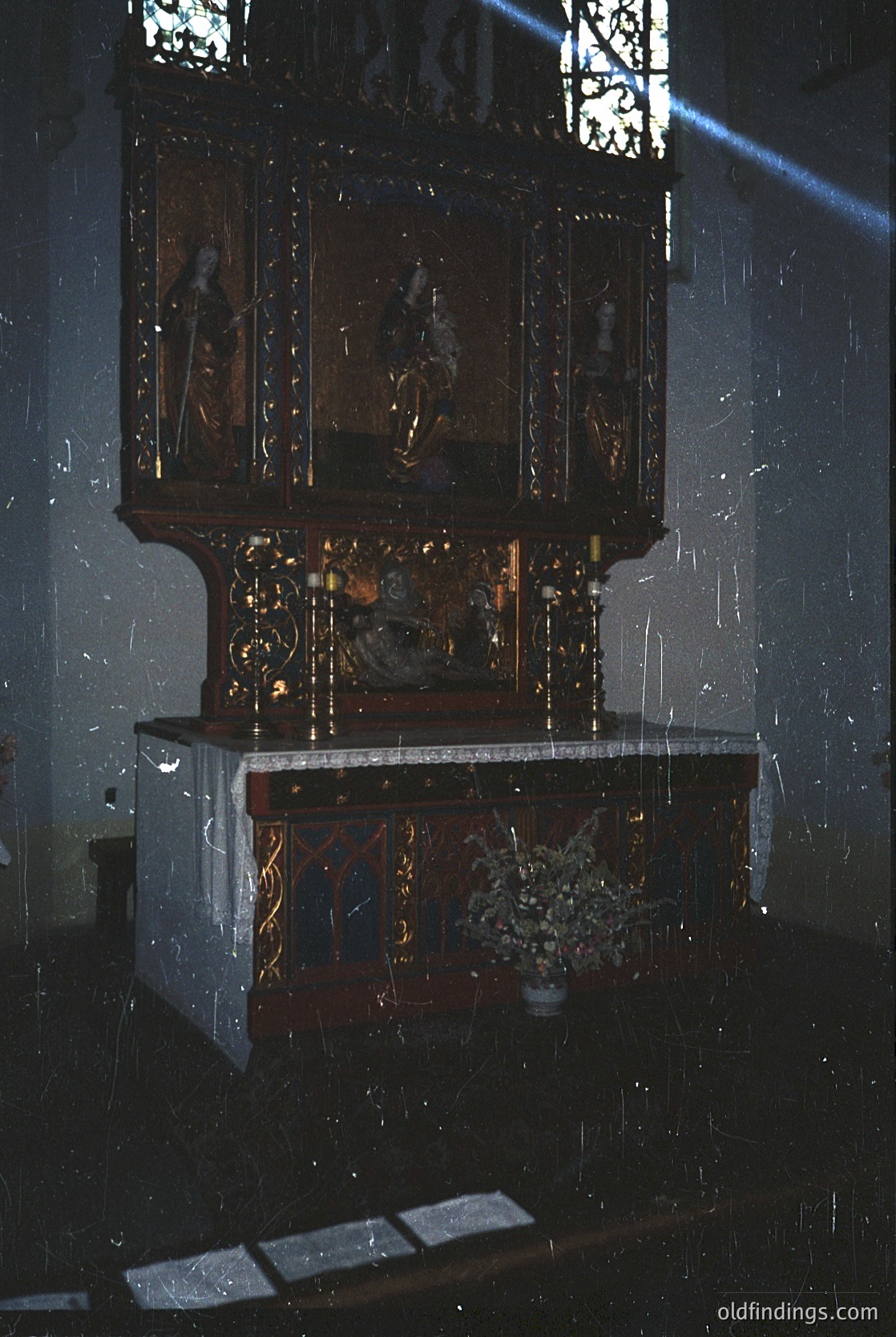 Ornate wooden iconostasis with religious iconography, featuring five painted panels depicting saints and biblical scenes. Marble altar table with floral arrangement, likely Eastern Orthodox church interior. Gothic-style stained-glass window casting colored light.
