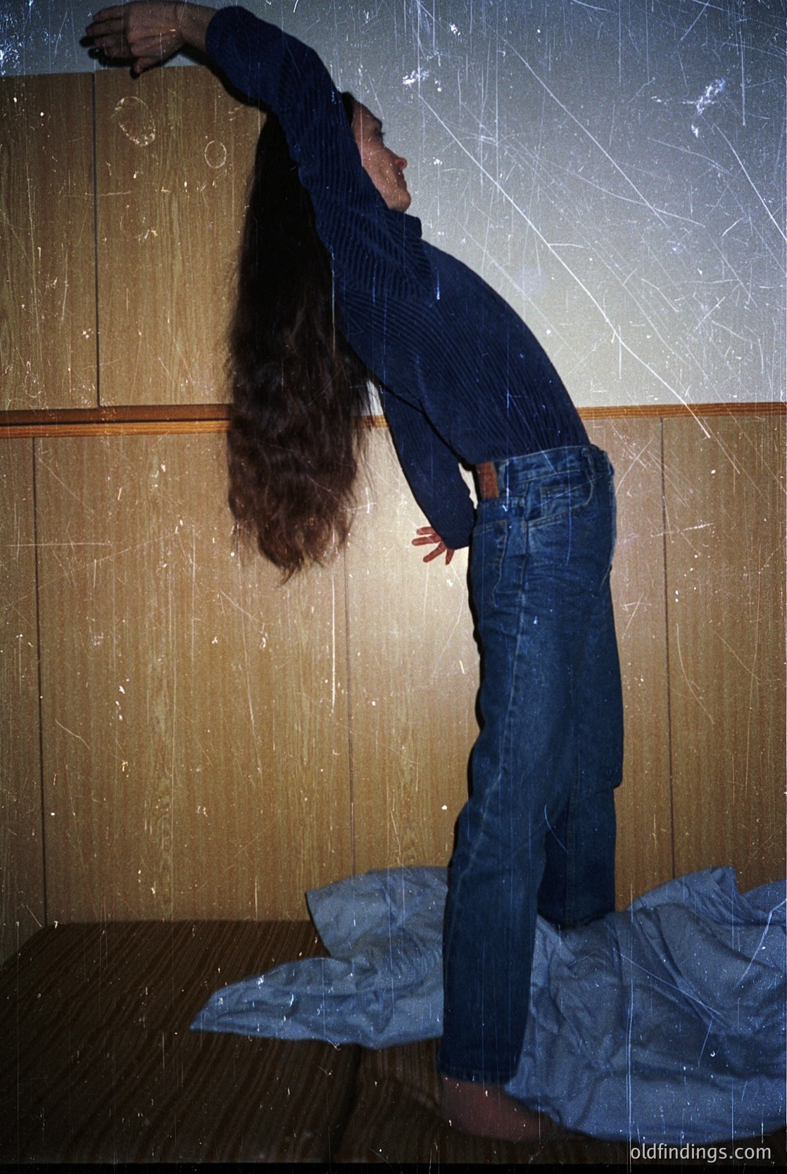 Portrait of a person mid-stretch against a textured wall, likely a 1970s interior. Dark blue striped sweater, high-waisted jeans, and long hair framing the pose. Minimalist composition with a single folded blanket on the floor.