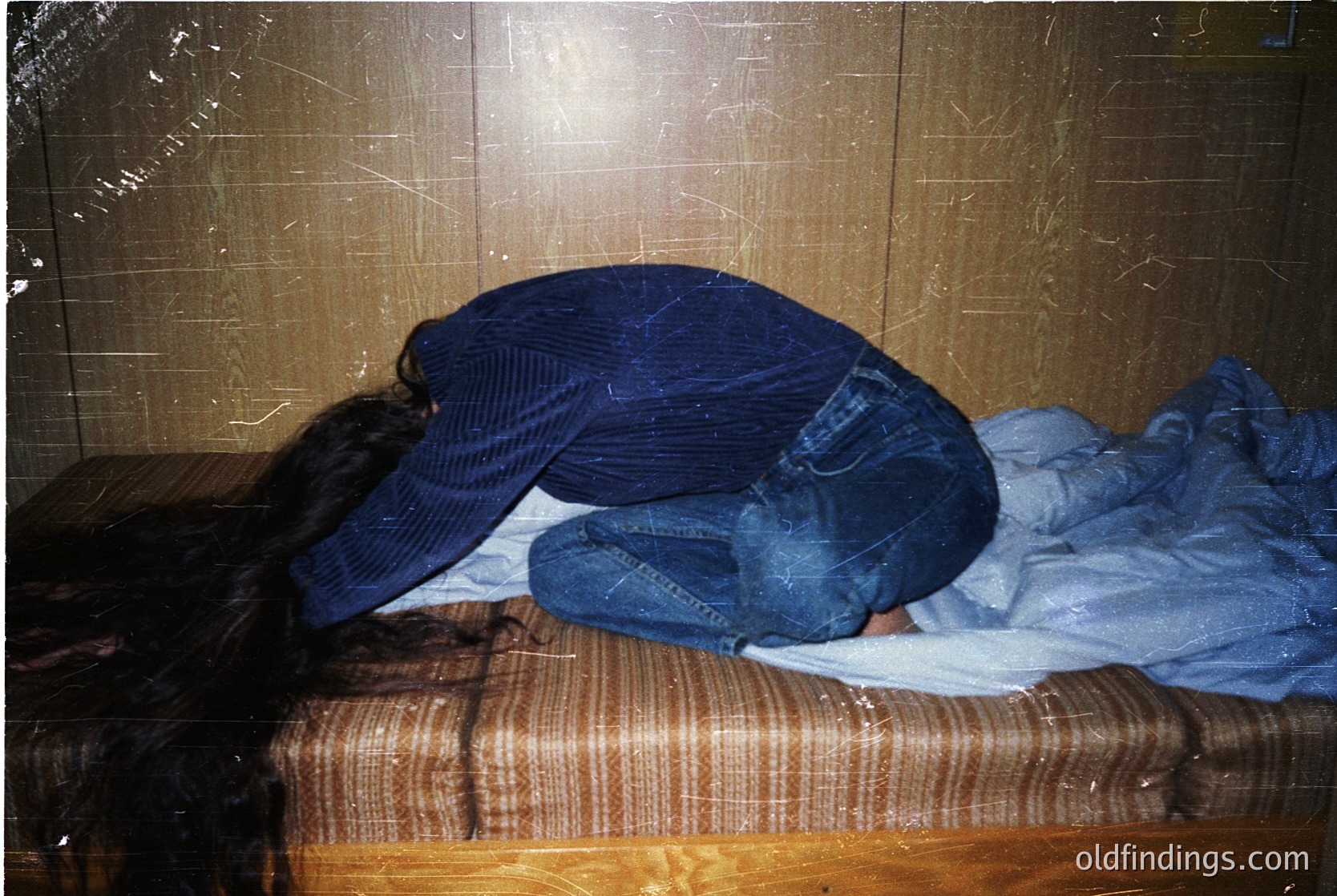 Person resting head on a wooden bench in an indoor setting, likely a public transport station or waiting area. The worn, striped shirt and jeans suggest mid-20th century fashion. Tiled wall and scratched metal ceiling indicate utilitarian design.