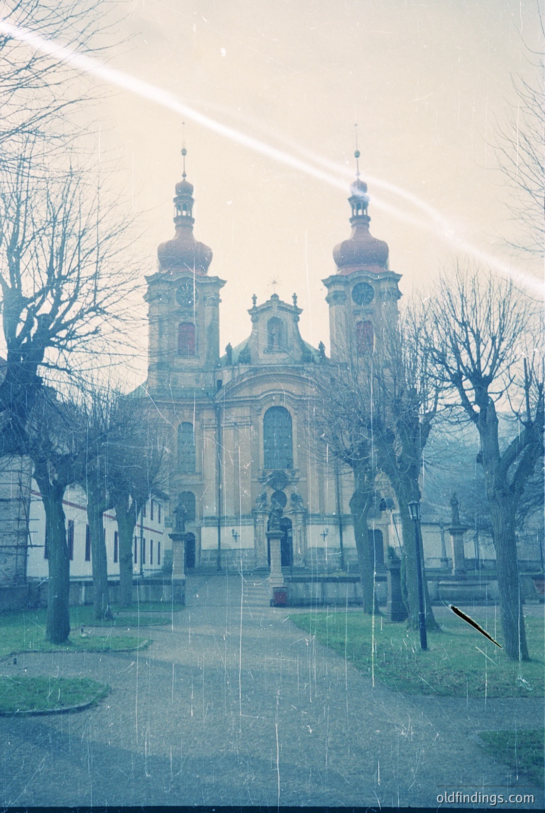 Baroque-style church with twin domed towers and ornate façade, framed by bare winter trees. Symmetrical clock placement on central pediment. Courtyard with gravel paths and sculpted lamp posts. Likely European, 18th-century architecture.