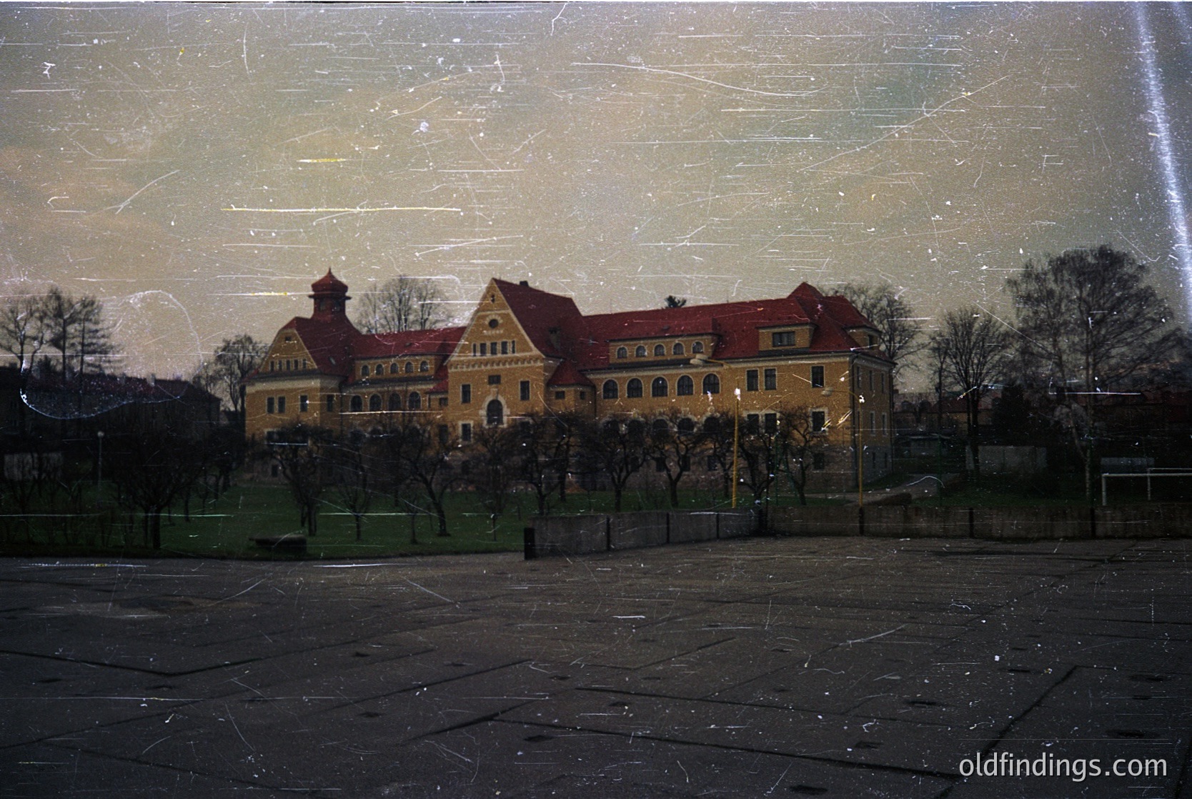 Vintage sepia-toned photograph of a grand, symmetrical **Art Nouveau-style building** with red-tiled roofs and arched windows. The structure features a central tower and a symmetrical façade, framed by leafless trees in a landscaped courtyard. Likely **early 20th century**, possibly **Europe**.