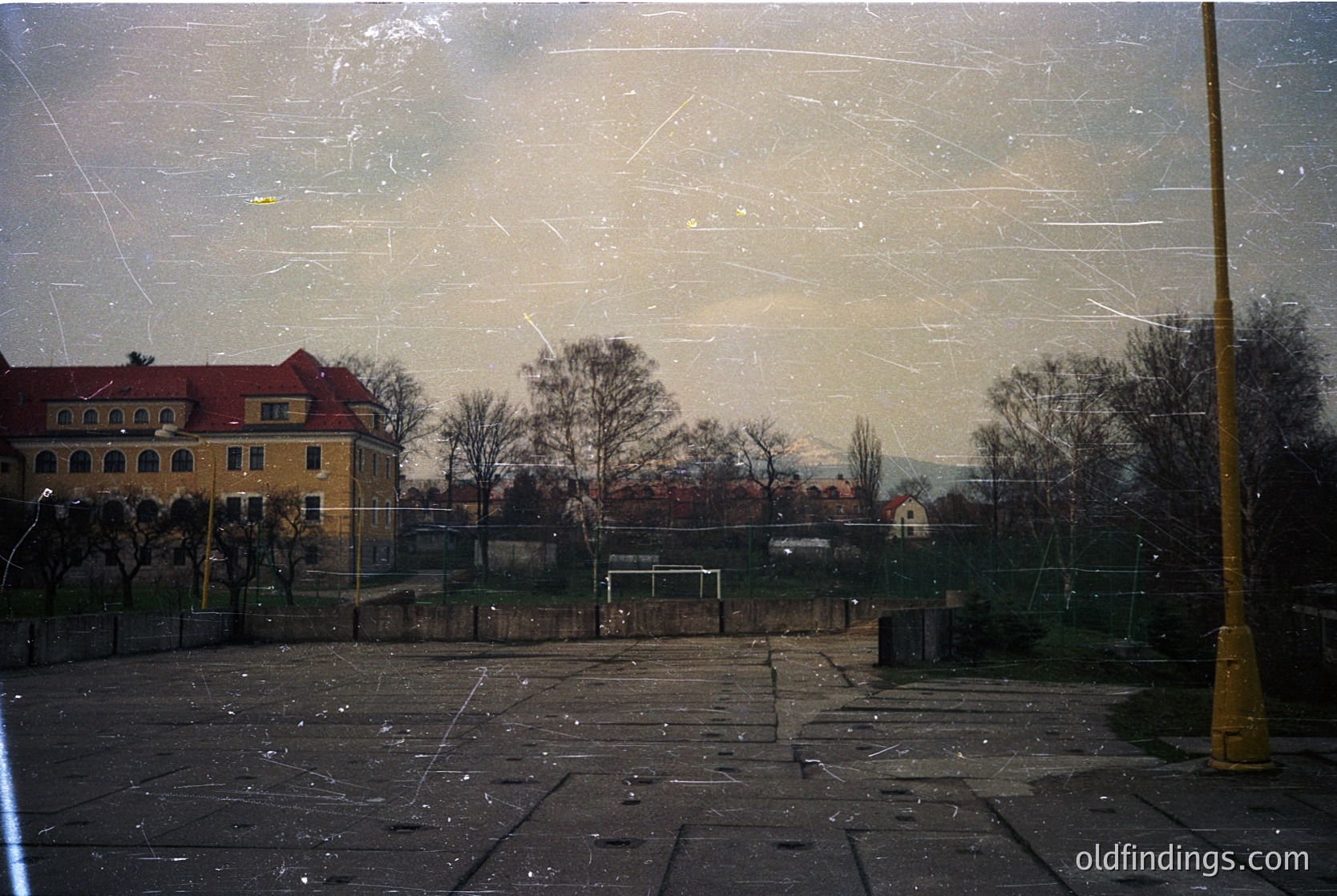 Vintage urban scene through a fogged window, showing a yellow brick building with arched windows and a red-tiled roof. Empty courtyard with bare trees, soccer goal, and cracked pavement. Overcast skies and muted autumnal tones suggest late 20th-century European architecture.