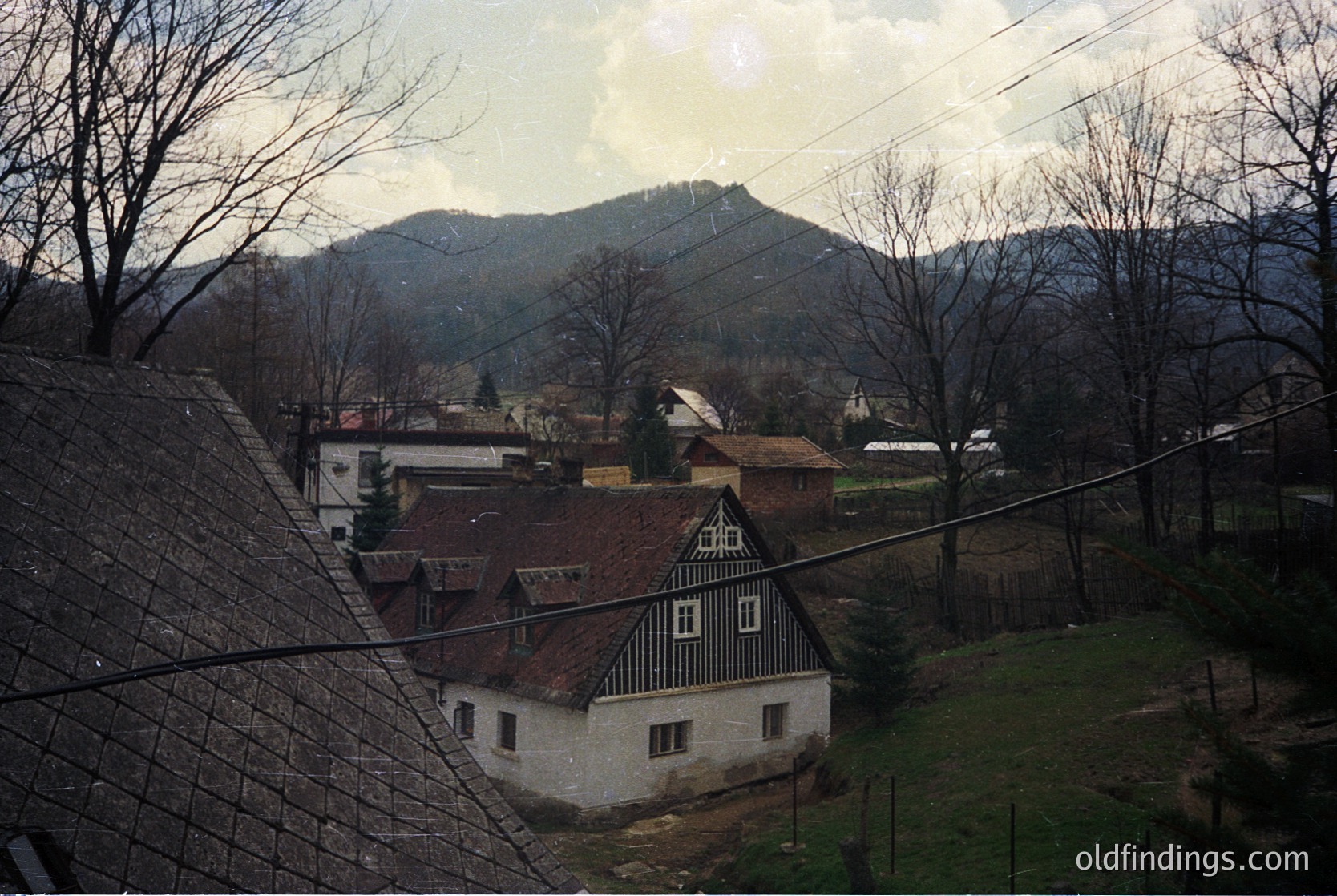 Rural village scene featuring a two-story timber-framed house with a steep gable roof, surrounded by leafless trees and rolling hills. Overhead power lines and a fence frame the foreground. Likely Eastern European countryside, mid-20th century.