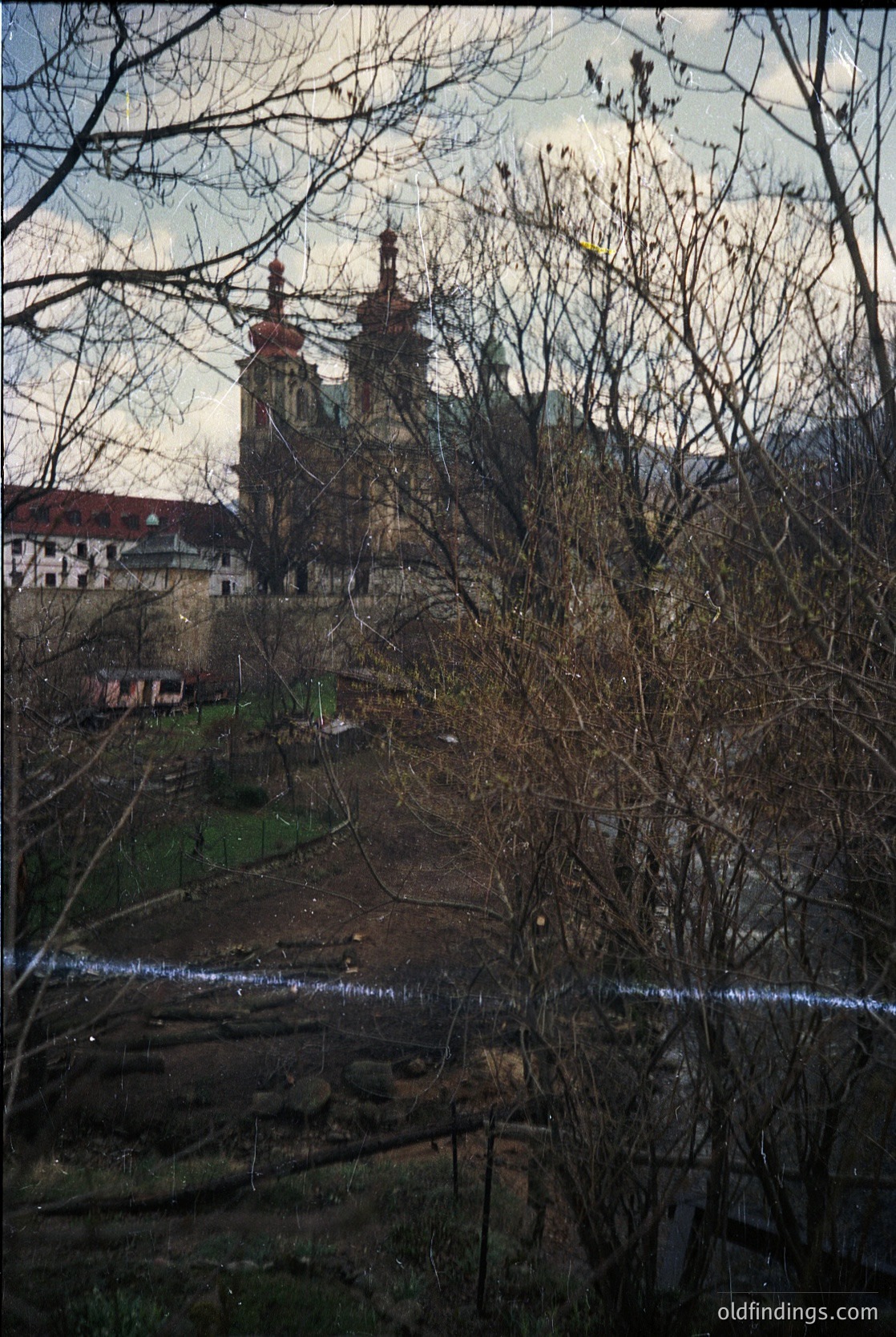 Baroque-style church with twin towers and ornate façade framed by winter trees. Urban landscape with residential buildings in background. Snow-covered ground suggests late winter or early spring. Likely European setting, possibly Central/Eastern Europe.
