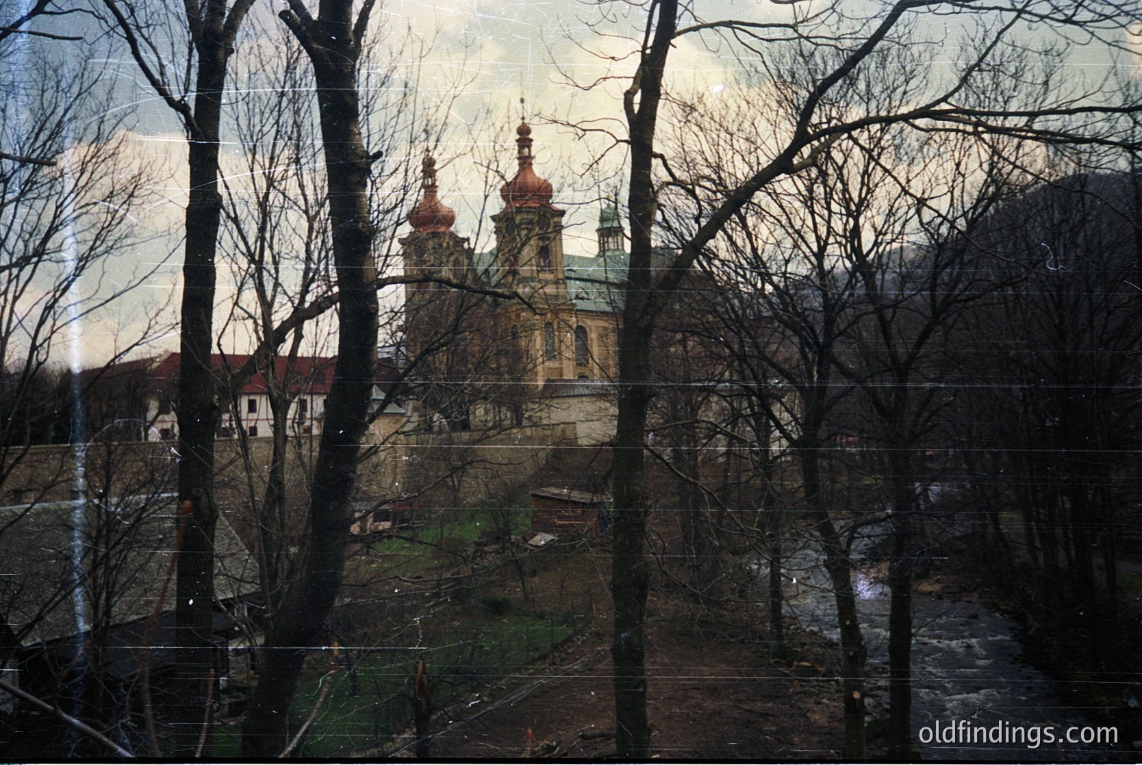 Baroque-style church with twin golden domes and green-roofed spires framed by bare winter trees. Overcast sky enhances muted tones. Likely Eastern European architecture, possibly or , 18th–19th century.
