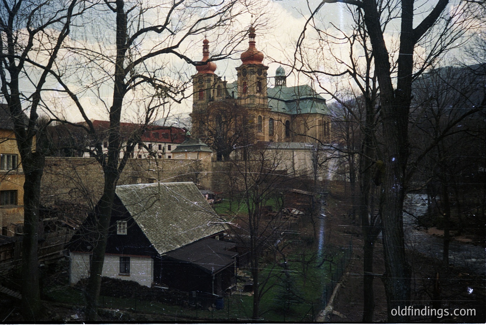 Baroque-style church with twin towers and domed roof framed by winter trees, likely Eastern European. Rustic wooden houses and muted urban landscape in foreground. *(Note: Exact location/time indeterminate but stylistic cues suggest 1800s Eastern Europe.)*