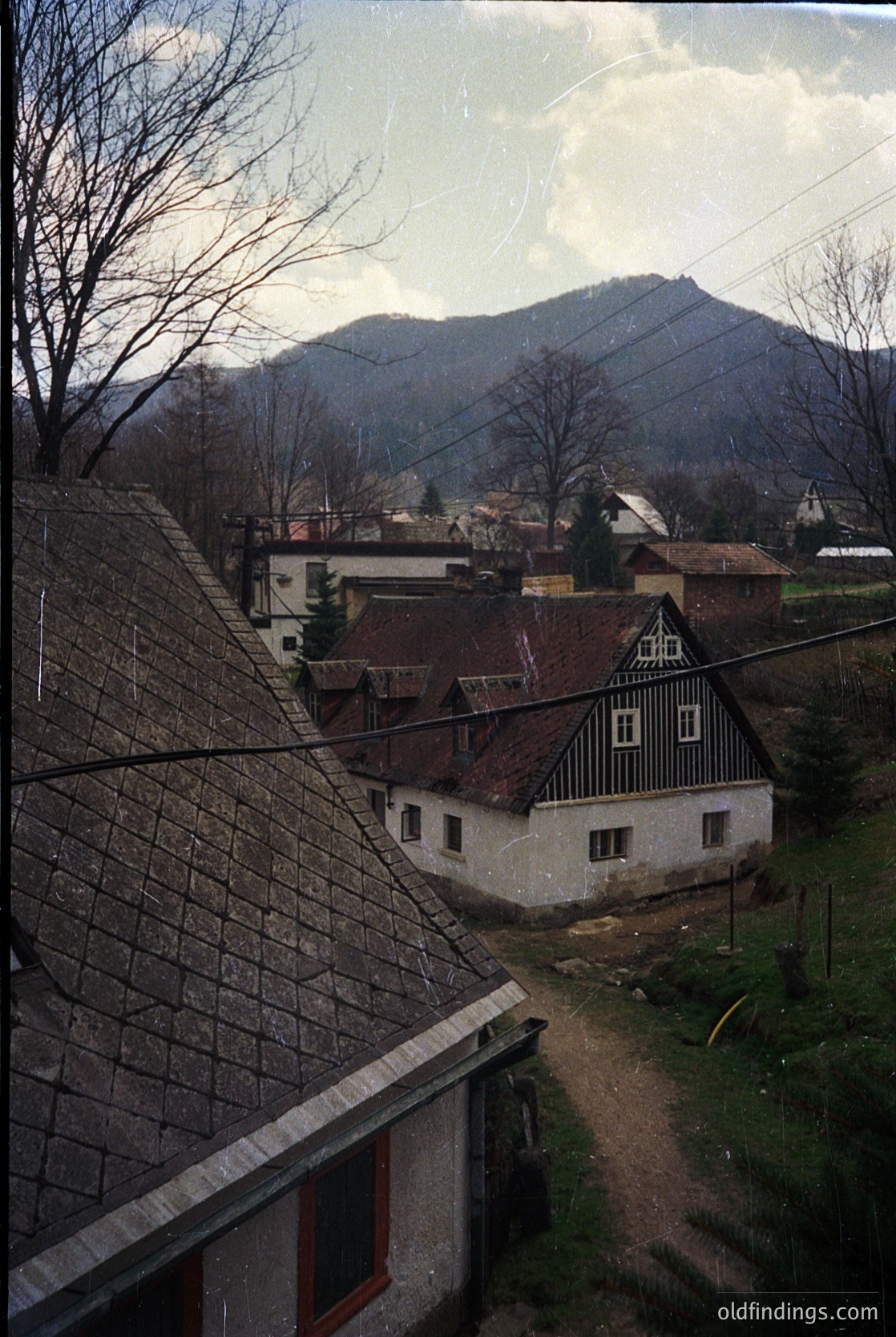 Rustic alpine village with timber-framed houses and stone roofs, framed by bare winter trees. Snow-capped mountains loom in the background under a cloudy sky. Likely Eastern European, mid-20th century.