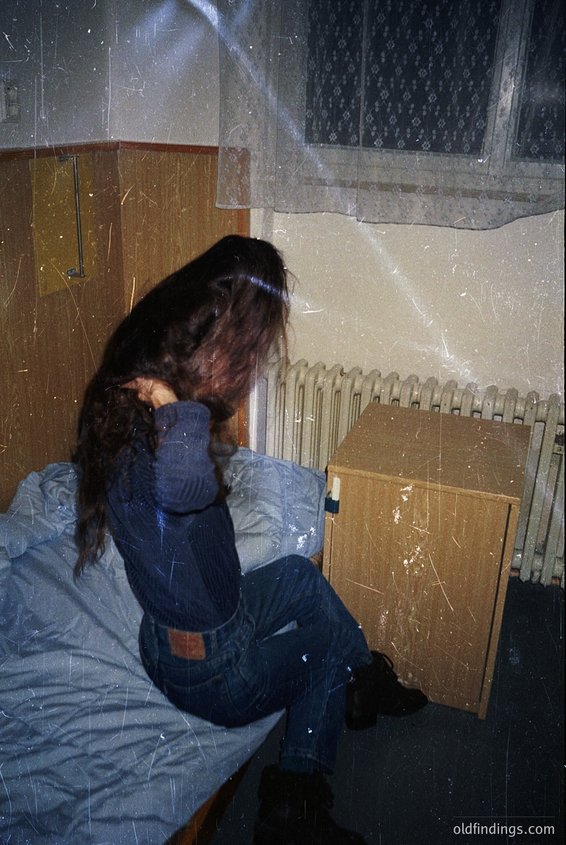 Vintage-style interior shot of a person sitting on a bed with a radiator beside them. The individual wears a long-sleeve shirt, jeans, and boots, with long hair. The room features peeling wallpaper and a window with frosted glass. Likely mid-20th century domestic setting.