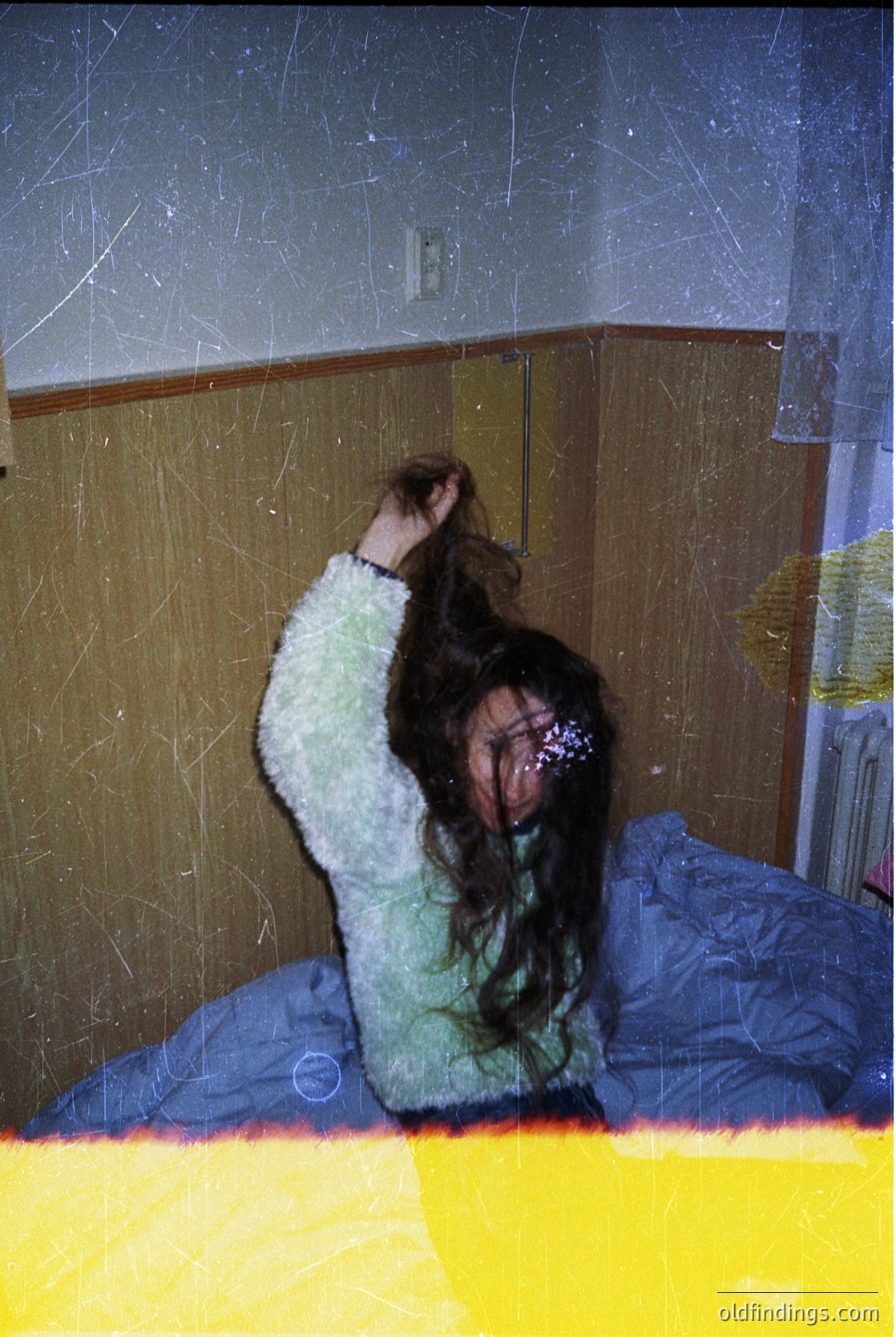 Vintage candid of a person in a green sweater and barrette, seated on a bed with a blue blanket. Interior features textured wallpaper and a wooden headboard. Likely mid-20th century domestic setting.