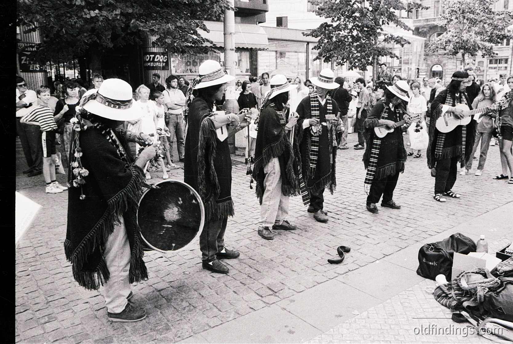 Street scene featuring a traditional folk band in traditional attire, likely Bulgarian. Musicians play accordions, drums, and stringed instruments while wearing wide-brimmed hats and embroidered vests. Crowd gathers around, suggesting a public festival or cultural event. Urban setting with cobblestone pavement and storefronts in background.