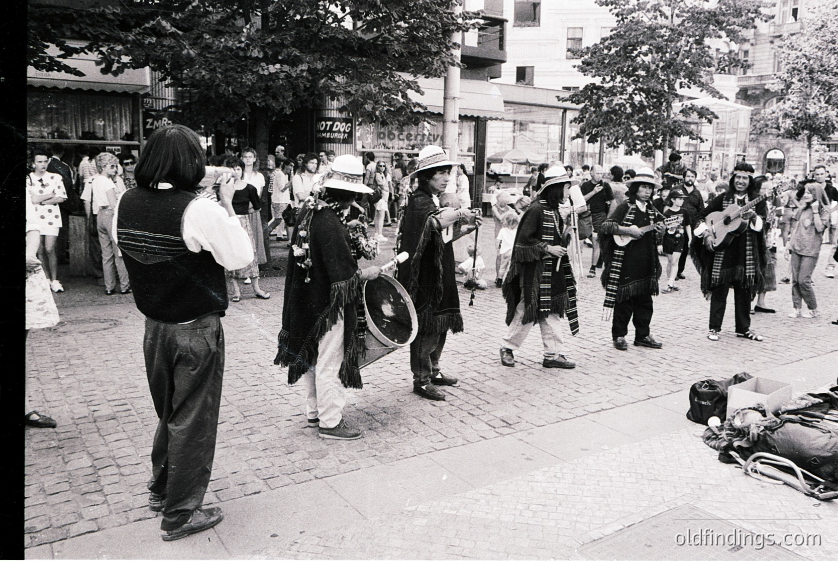 Street performance featuring traditional musicians in folk attire, likely Eastern European. Group plays accordion, drums, and string instruments in a cobblestone plaza. Crowd gathers, some recording or photographing. Urban setting with mid-rise buildings and storefronts in background.