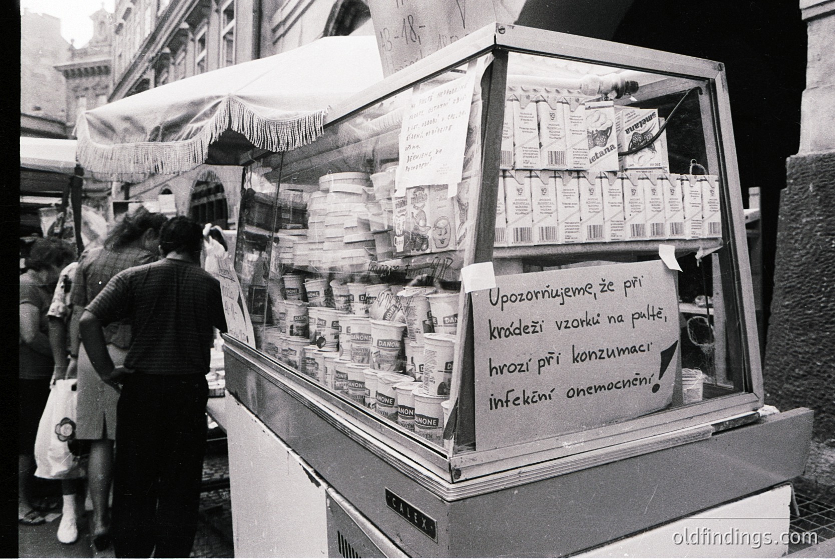 Street-side vending cart in black-and-white, displaying packaged dairy products (milk, yogurt) with handwritten warning in Czech: *"Upozorněme že při kradeži vzorků na paletě, hrozí při konzumaci infekční onemocnění!"* (Warning: theft of samples from pallets poses infection risk if consumed). Urban setting, likely 1970s–1980s Eastern Europe.