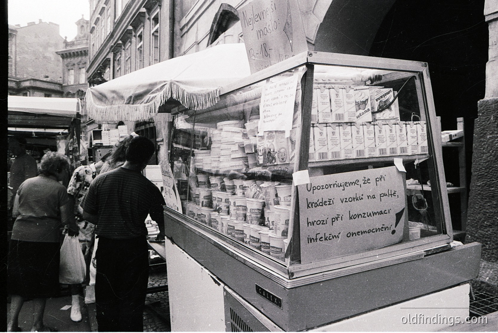 Street-side ice cream stall in urban setting, likely Eastern Europe, 1960s–70s. Handwritten sign warns of hygiene risks ("Upozorňujeme..."). Packaged ice cream and drinks displayed in glass case. Crowd in mid-20th-century attire.