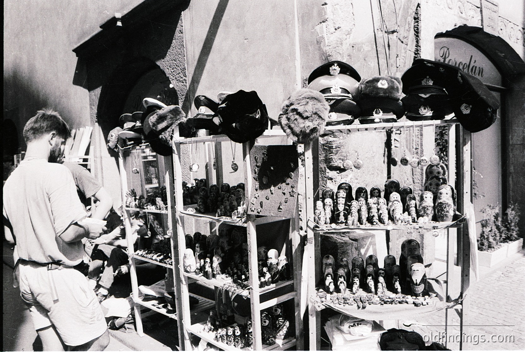 Vintage street vendor stall showcasing military hats and stuffed animal figurines in glass display cases, likely mid-20th century. Note the "Borsalino" branded hat on the right. Urban setting with visible architectural details like exposed brick.