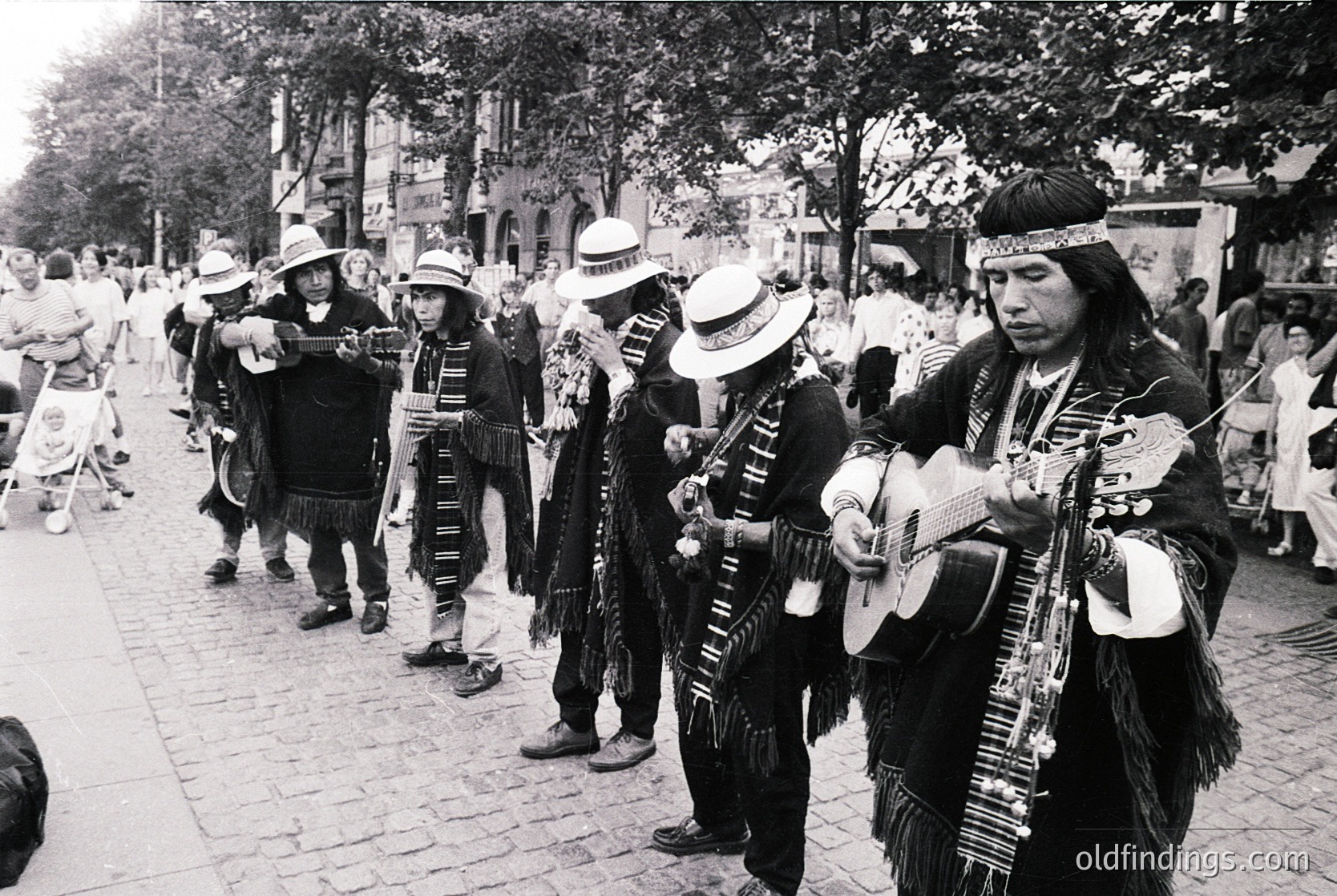 Street performance featuring traditional Andean musicians in elaborate, fringed ponchos and wide-brimmed hats, playing acoustic guitars and panpipes. Crowd and urban backdrop suggest a public square or plaza. Likely or Latin America cultural festival.
