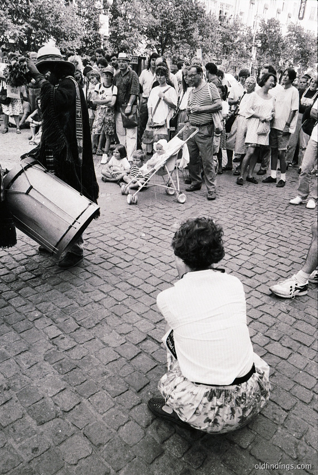 Street musician playing a large frame drum beside a seated woman in floral dress, 1970s urban setting. Crowd of casually dressed people observing. Cobblestone pavement and greenery in background.