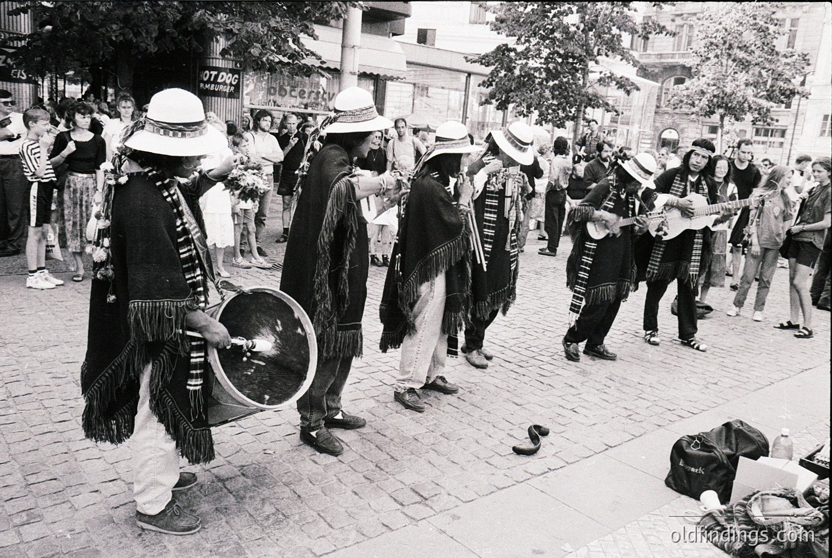 Street performance featuring traditional musicians in folk attire—wide-brimmed hats, layered vests, and fringe—playing drums, accordions, and string instruments. Urban setting with cobblestone pavement, spectators, and visible storefronts ( ).