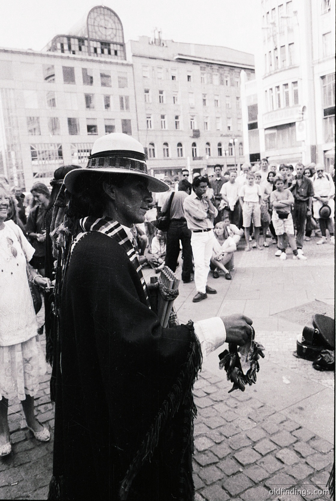 Street musician in traditional attire plays accordion to a gathered crowd in an urban plaza. Architectural details include mid-century concrete buildings with arched windows. Crowd wears 1960s-70s fashion—wide trousers, jackets, and hats.