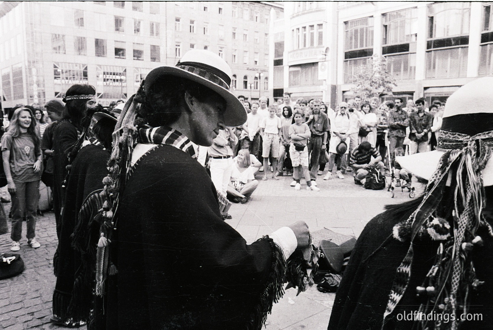 Crowded urban street scene featuring Indigenous performers in traditional regalia, likely during a cultural festival or protest. Cowboy hats, feathered vests, and face paint distinguish the group. Surrounding spectators, some wearing peace symbols, suggest 1960s–1970s activism era. Urban architecture hints at a North American city.
