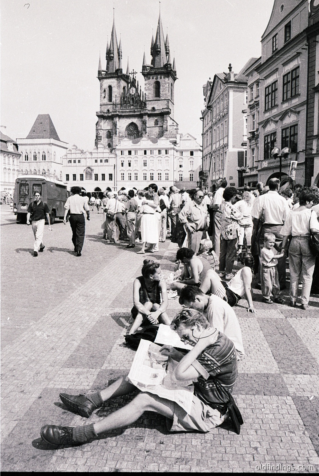 Crowd gathers in Prague’s Old Town Square, 1960s, with Gothic spires of Church of Our Lady before the Virgin Mary and St. James in background. Couple sits on cobblestones, while others stand or walk near historic buildings. Mid-century European street life captured.