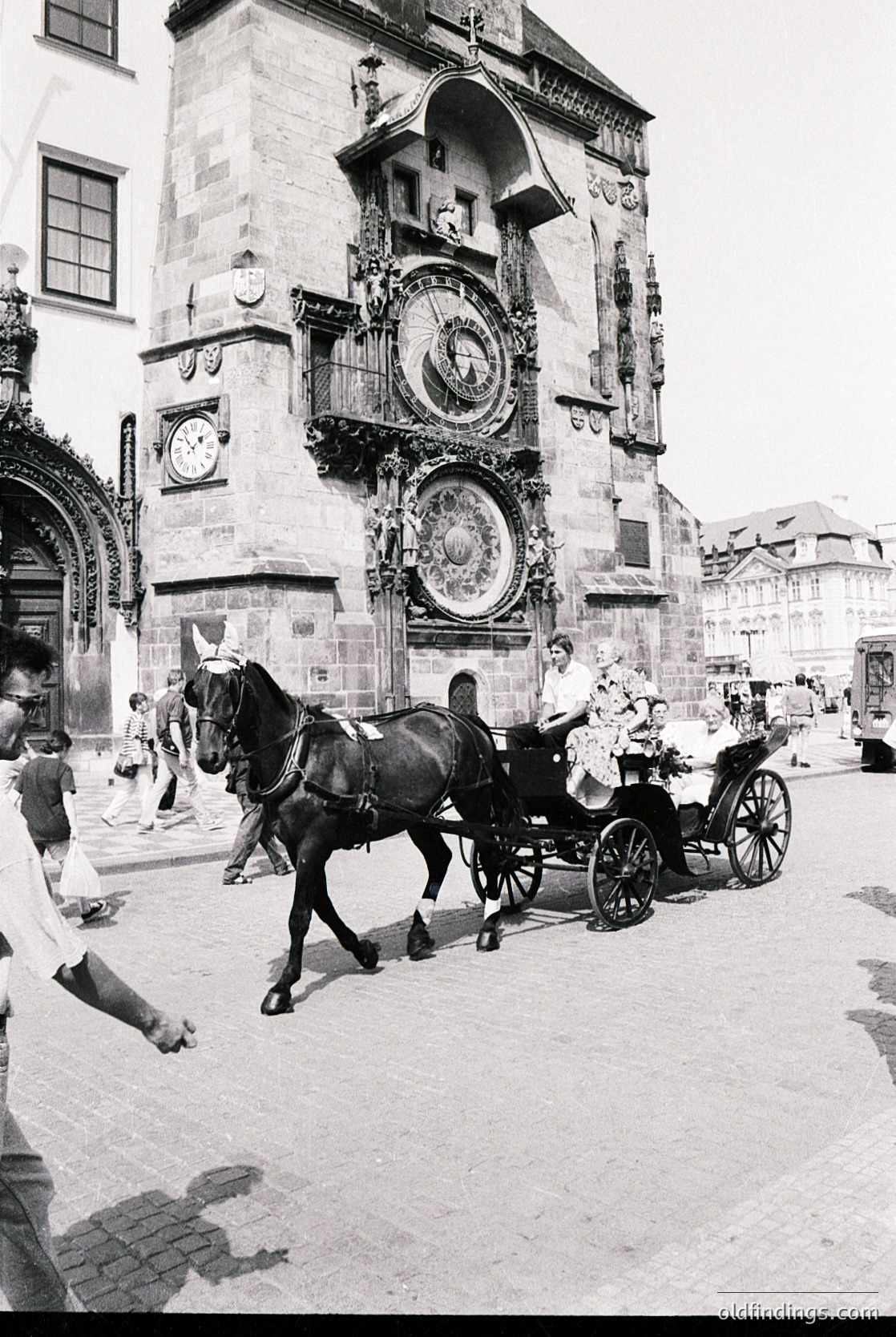 Historic horse-drawn carriage passing Prague’s Astronomical Clock tower (Orloj), 15th-century Gothic architecture with medieval astronomical dials. Cobblestone street bustling with pedestrians in 1950s-era attire. Iconic