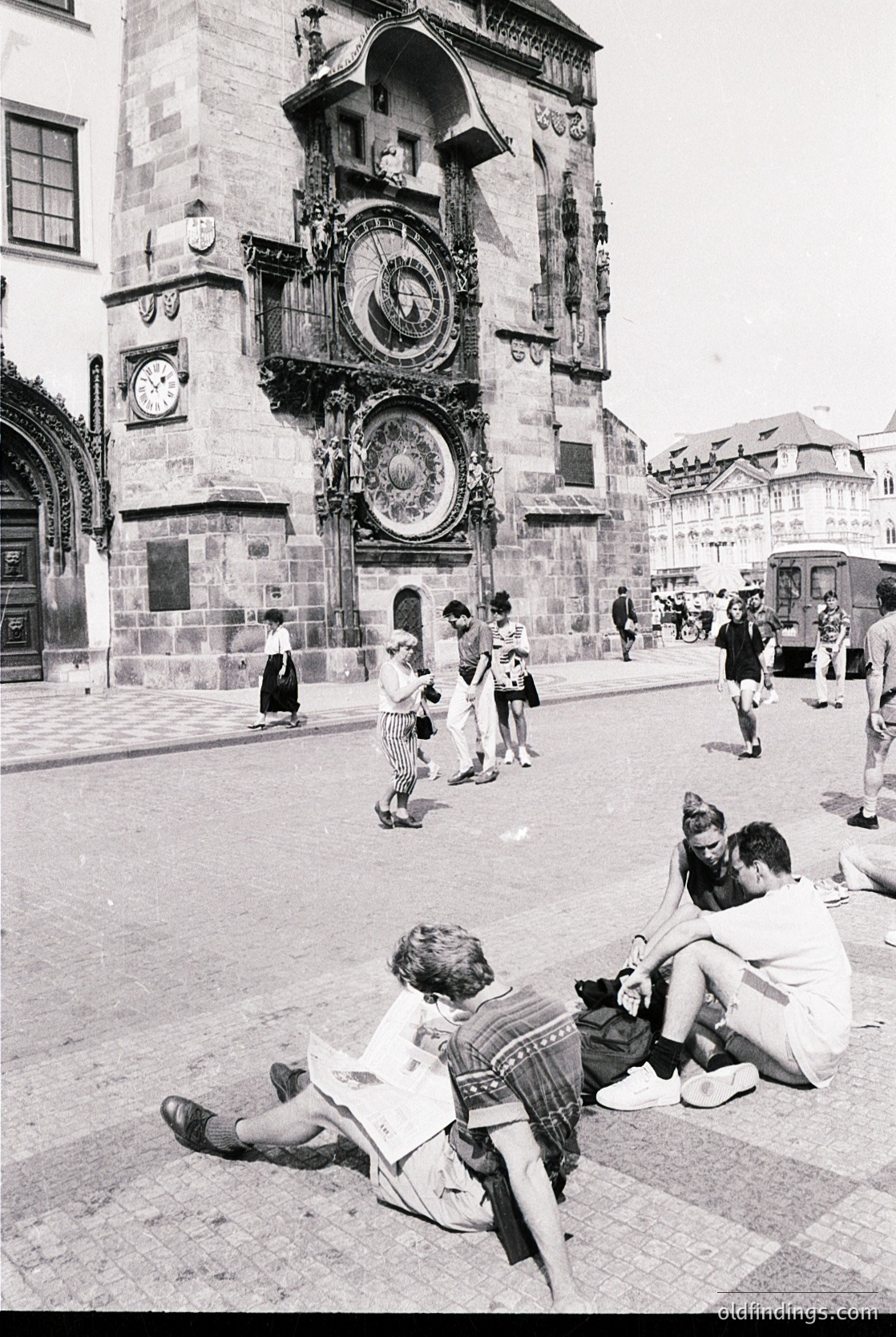 Mid-20th century black-and-white shot of Prague’s Astronomical Clock tower () with medieval Gothic architecture (). Tourists and locals gather on Old Town Square (), with two children sitting on pavement in foreground. Mid-century fashion () and urban European street life evident.