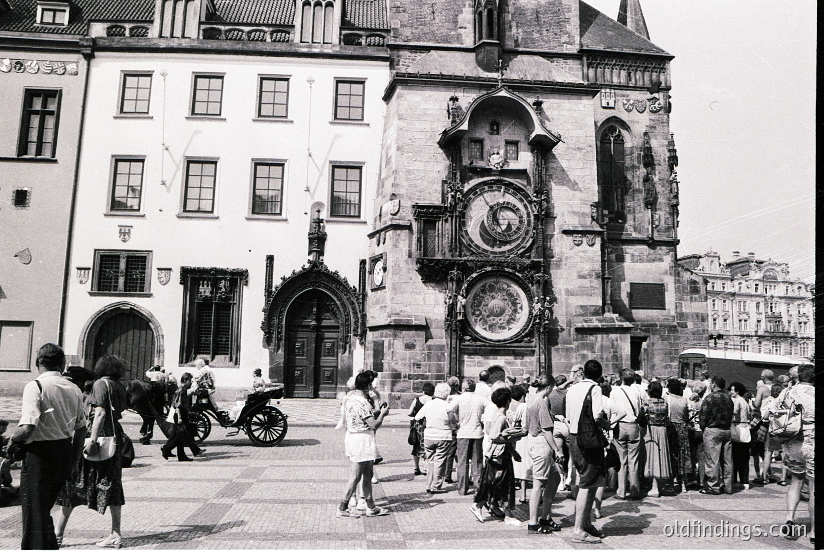 Historic Prague square featuring the Astronomical Clock Tower (Orloj) with medieval architecture. Crowds gather around the ornate clock face, showcasing Gothic-style details. Mid-20th century attire suggests or era.