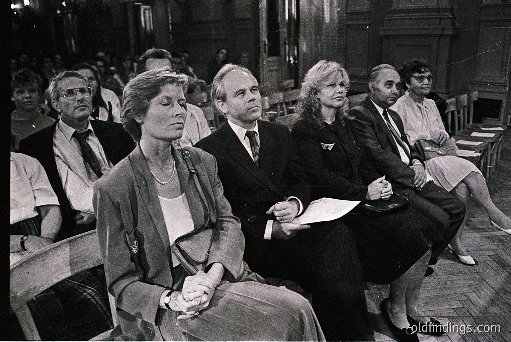 Black-and-white interior shot of seated attendees in a formal event, likely a 1970s–1980s conference or trial. Wooden pews and stone architecture suggest a courtroom or lecture hall. Attendees wear business attire: blazers, ties, and structured hairstyles. Central figures appear focused, with one woman holding papers.