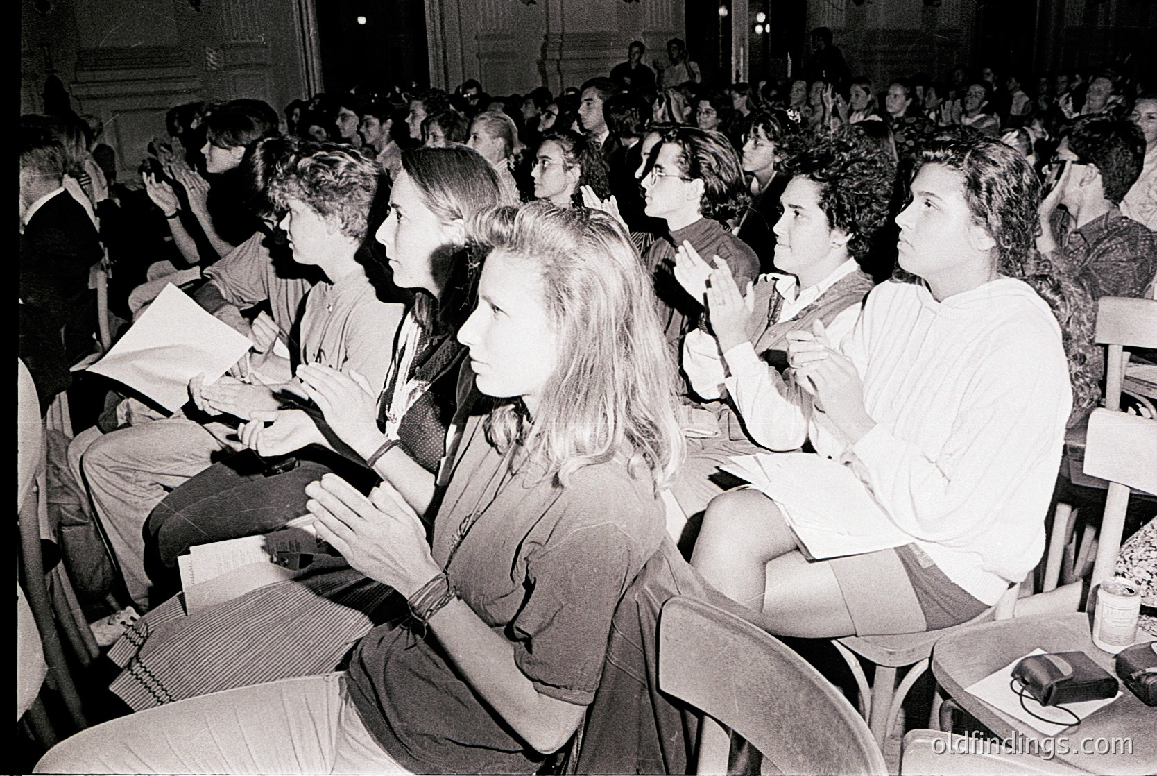 Black-and-white photo of a packed lecture hall, likely from the 1960s–1970s. Attendees, mostly young adults, sit attentively with notebooks and pens, facing a stage. Formal attire suggests an academic or cultural event. High ceilings and classical architecture hint at a university or concert hall setting.