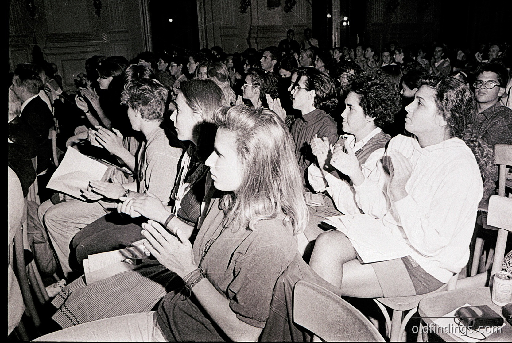 Crowded indoor lecture hall with seated students, likely 1960s–1970s. Wooden benches, formal attire (blazers, skirts), and classical architecture (columns, high ceilings) suggest an academic setting. Engaged audience with hands raised, indicating active participation.