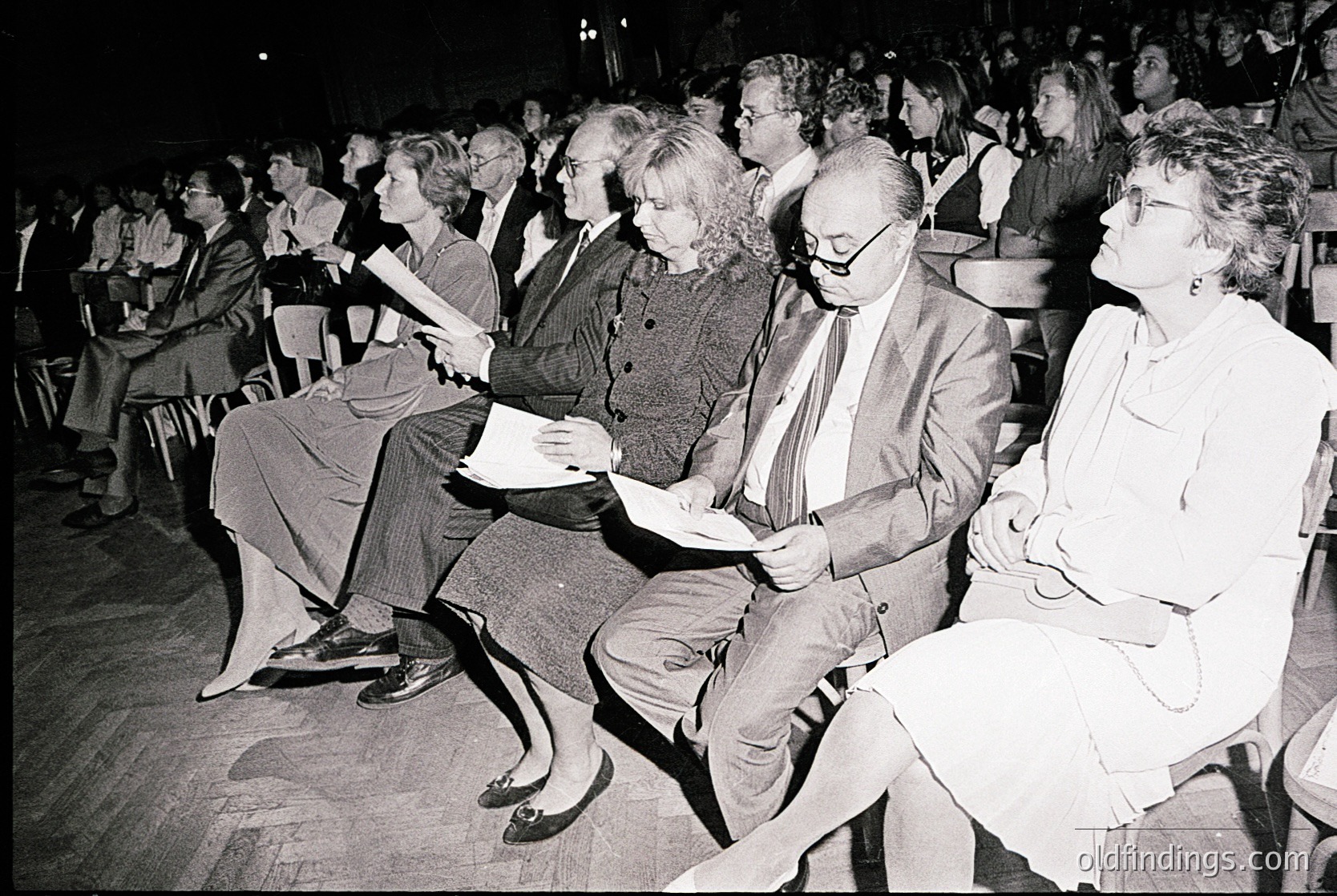 Black-and-white audience at a seated event, likely a lecture or performance, with attendees focused on the stage. Mid-20th century attire (1960s–1970s) suggests formal or intellectual gathering. Wooden floor and tiered seating indicate a university hall or cultural venue.