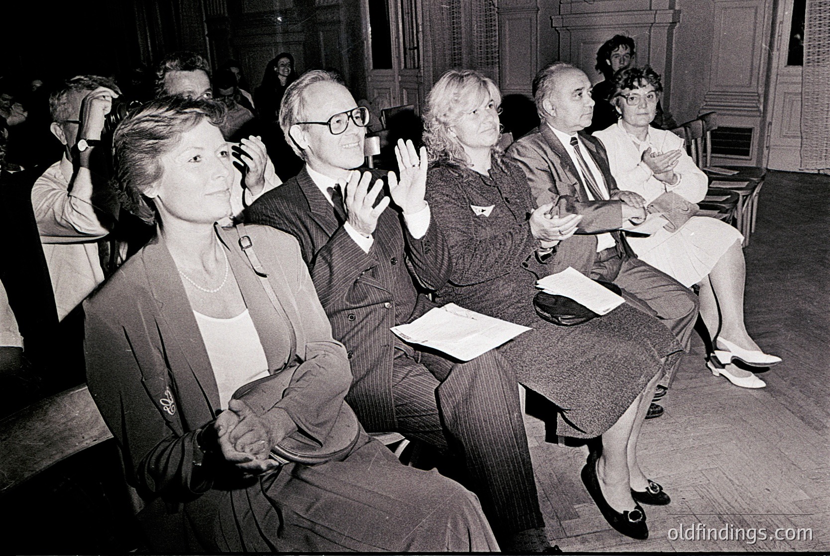 A black-and-white photo of an indoor audience in a formal setting, likely a 1970s conference or lecture. Six adults sit in wooden chairs, facing forward with attentive expressions. Men wear suits and glasses; women in blazers and dresses. A woman in the foreground raises her hand, possibly asking a question. Wood-paneled walls and a chandelier suggest an institutional or academic venue.
